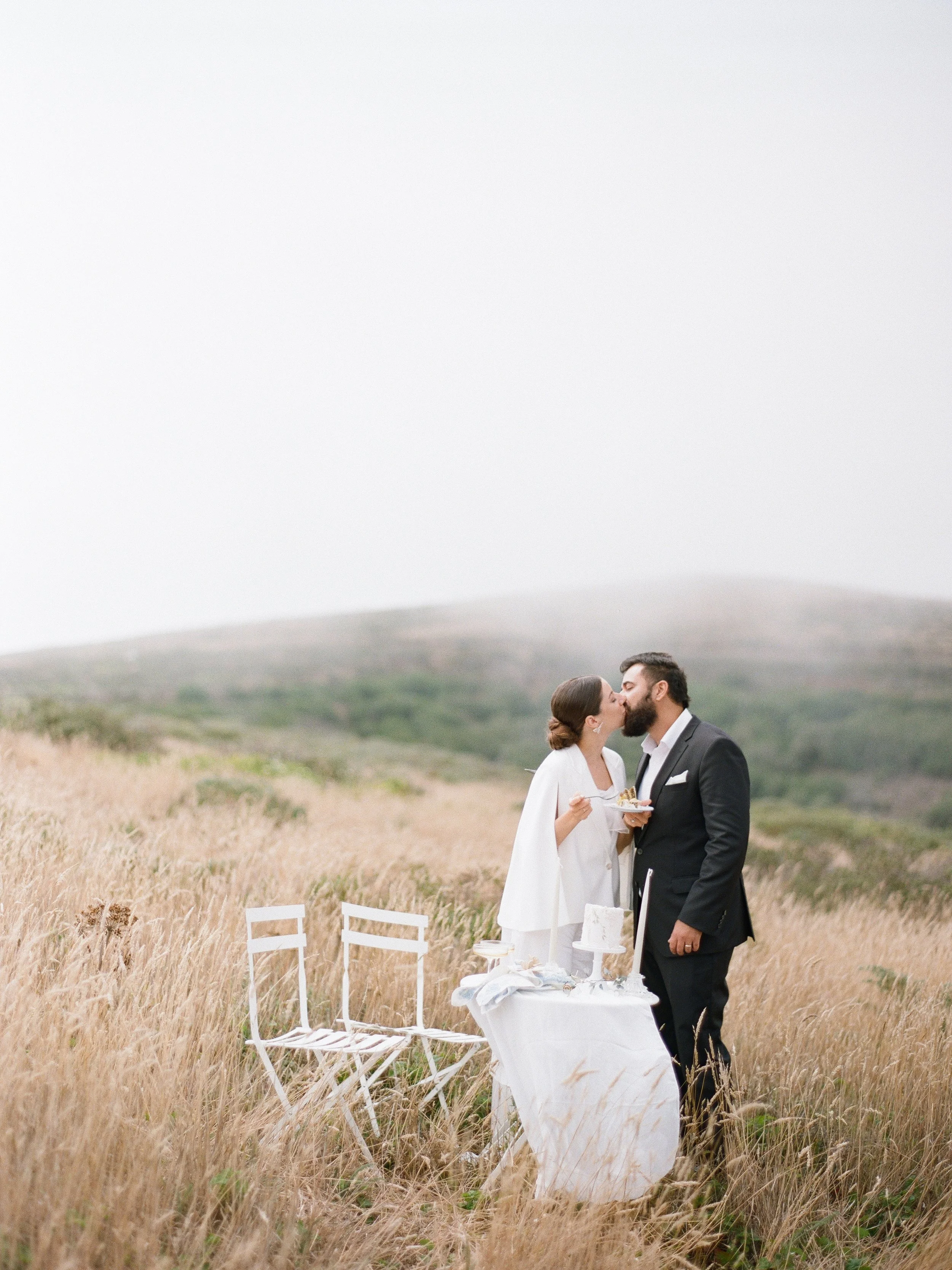Couple sharing a kiss in a foggy golden field at a California elopement, intimate table setup beside them, film photography by Radostina Boseva
