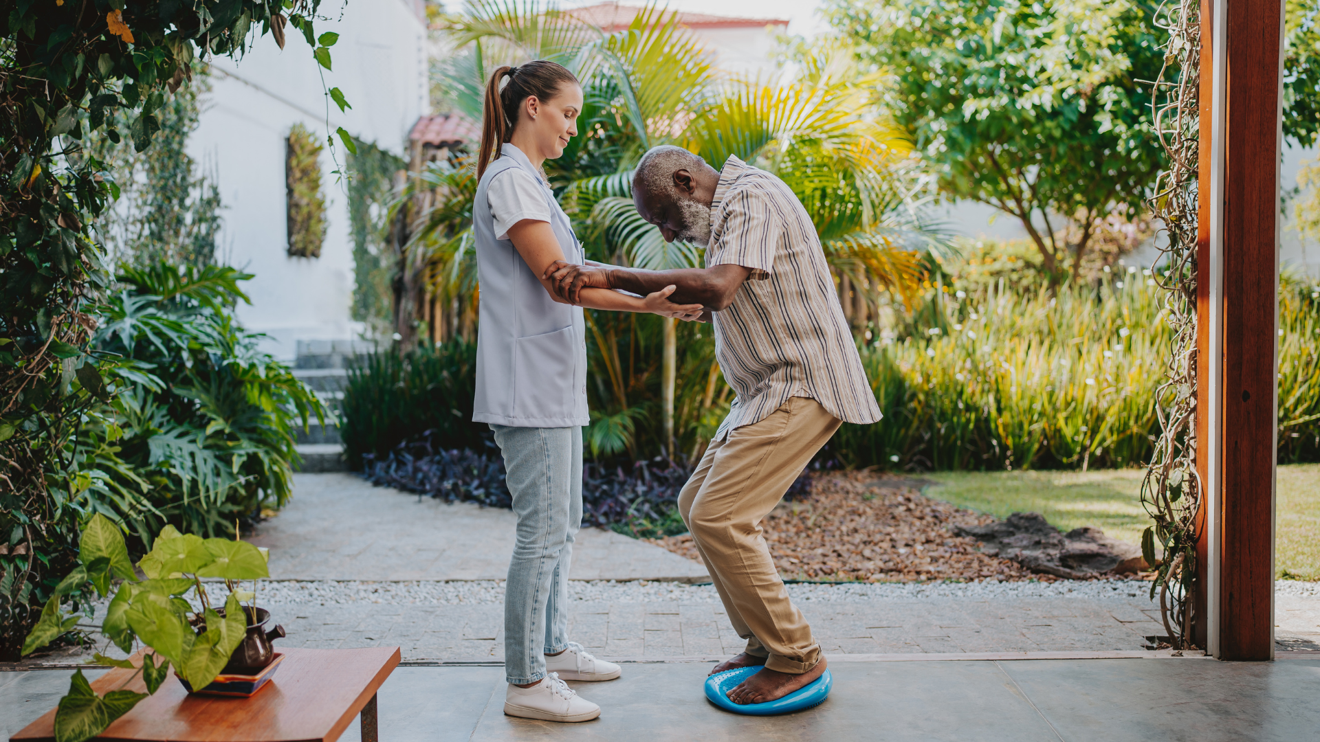 Physiotherapist supporting an older man during outdoor balance training on a wobble cushion in a home garden setting.