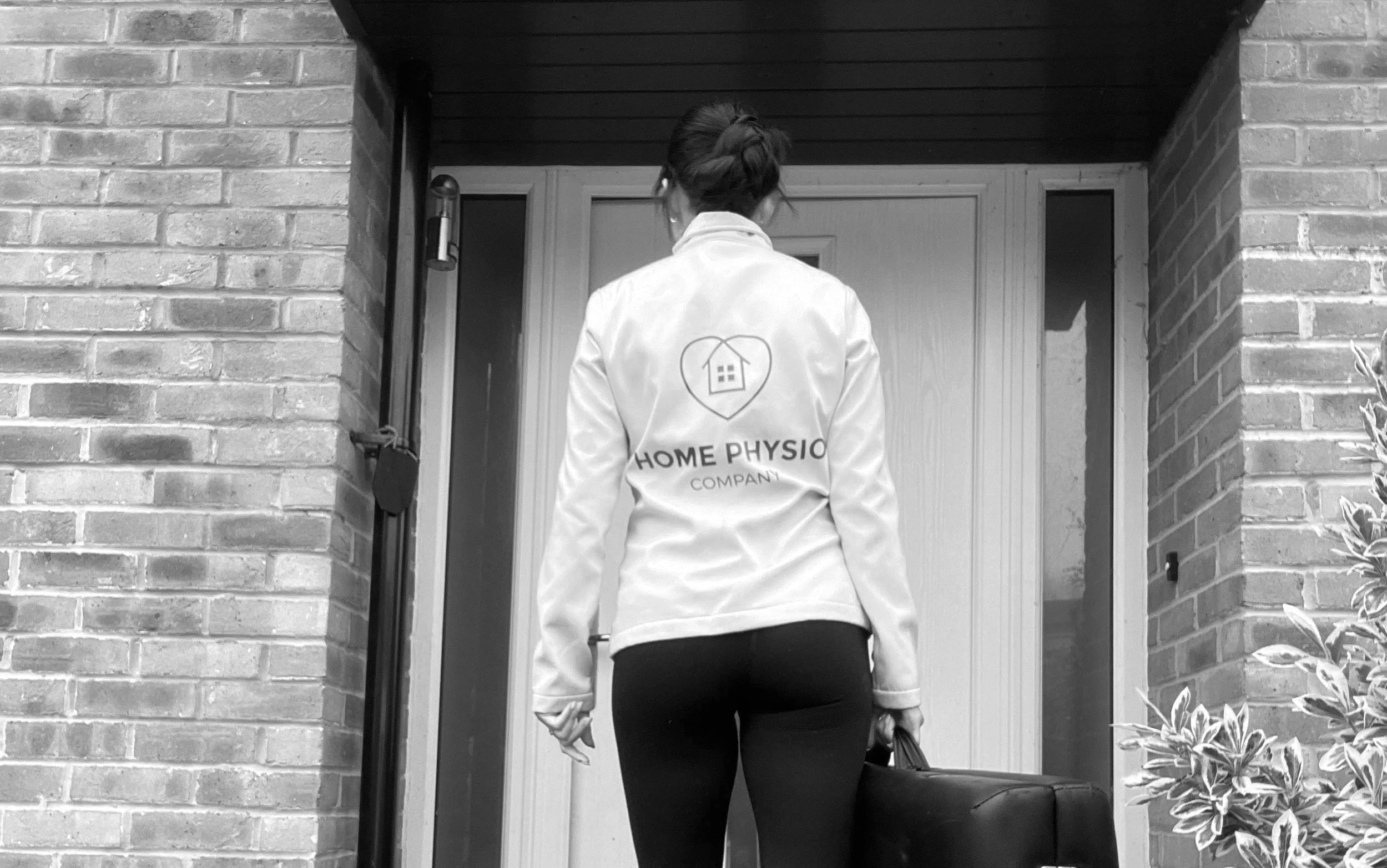 Female with dark hair and a top with Home Physio Company on the back holding a massage table approaching a front door