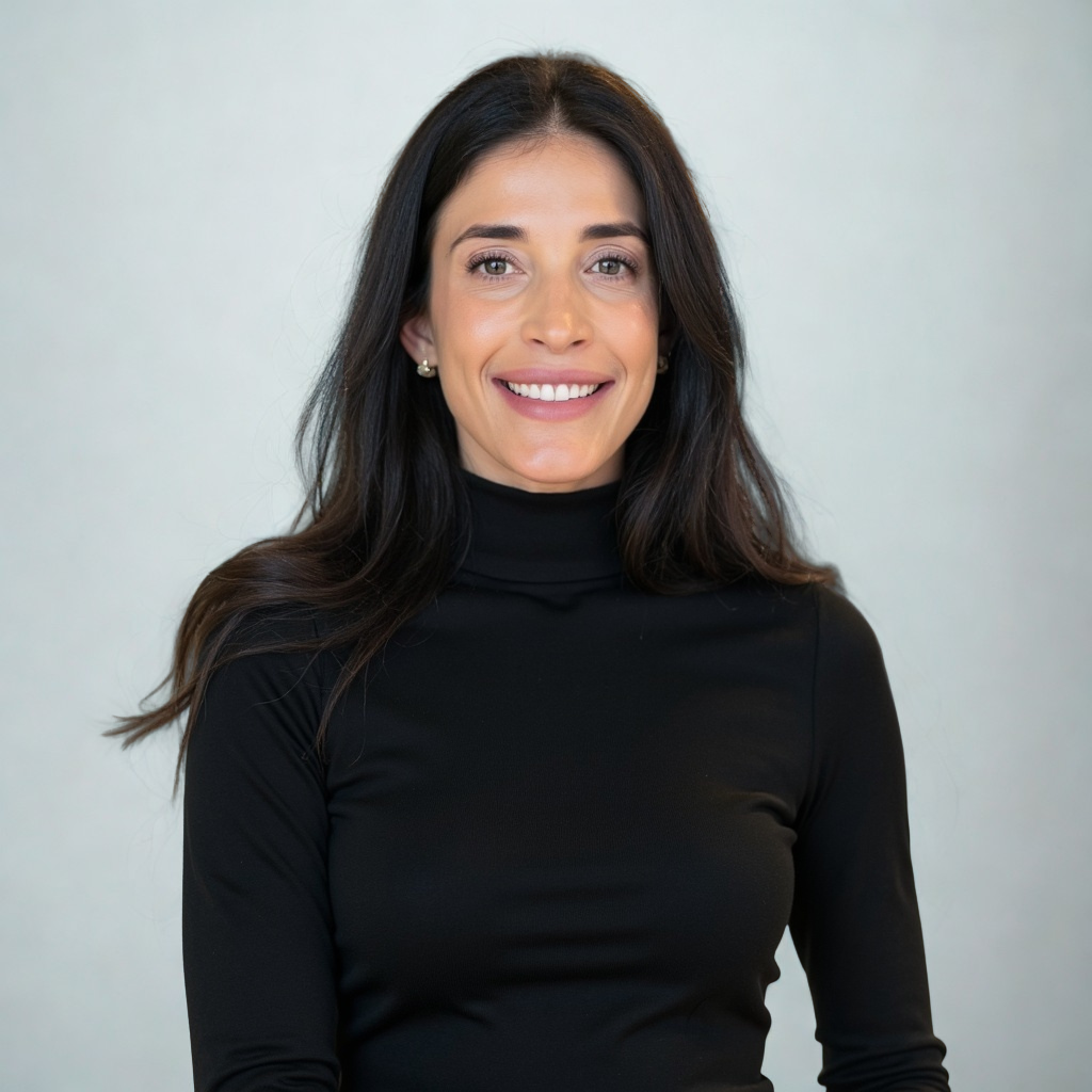A woman smiling, wearing a black turtleneck and pearl earrings, with long dark hair, standing against a light gray background.