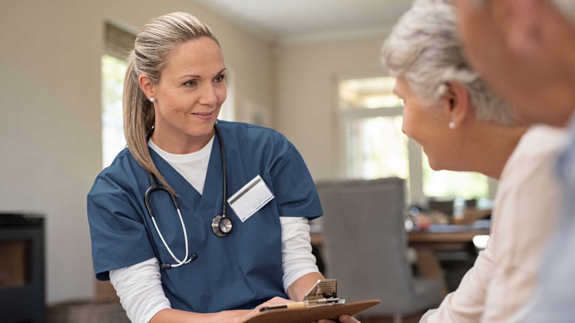 A nurse in blue scrubs and with a stethoscope around her neck talking to an elderly woman in a well-lit room.