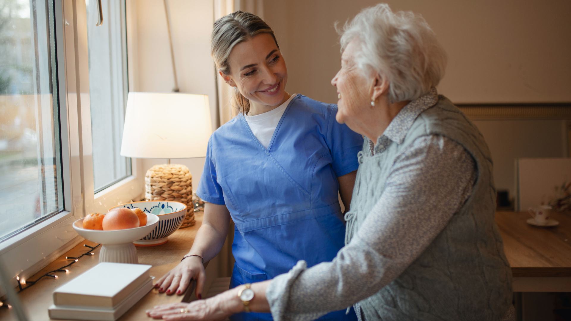 A young female caregiver in blue scrubs comforting an elderly woman in a cozy kitchen.
