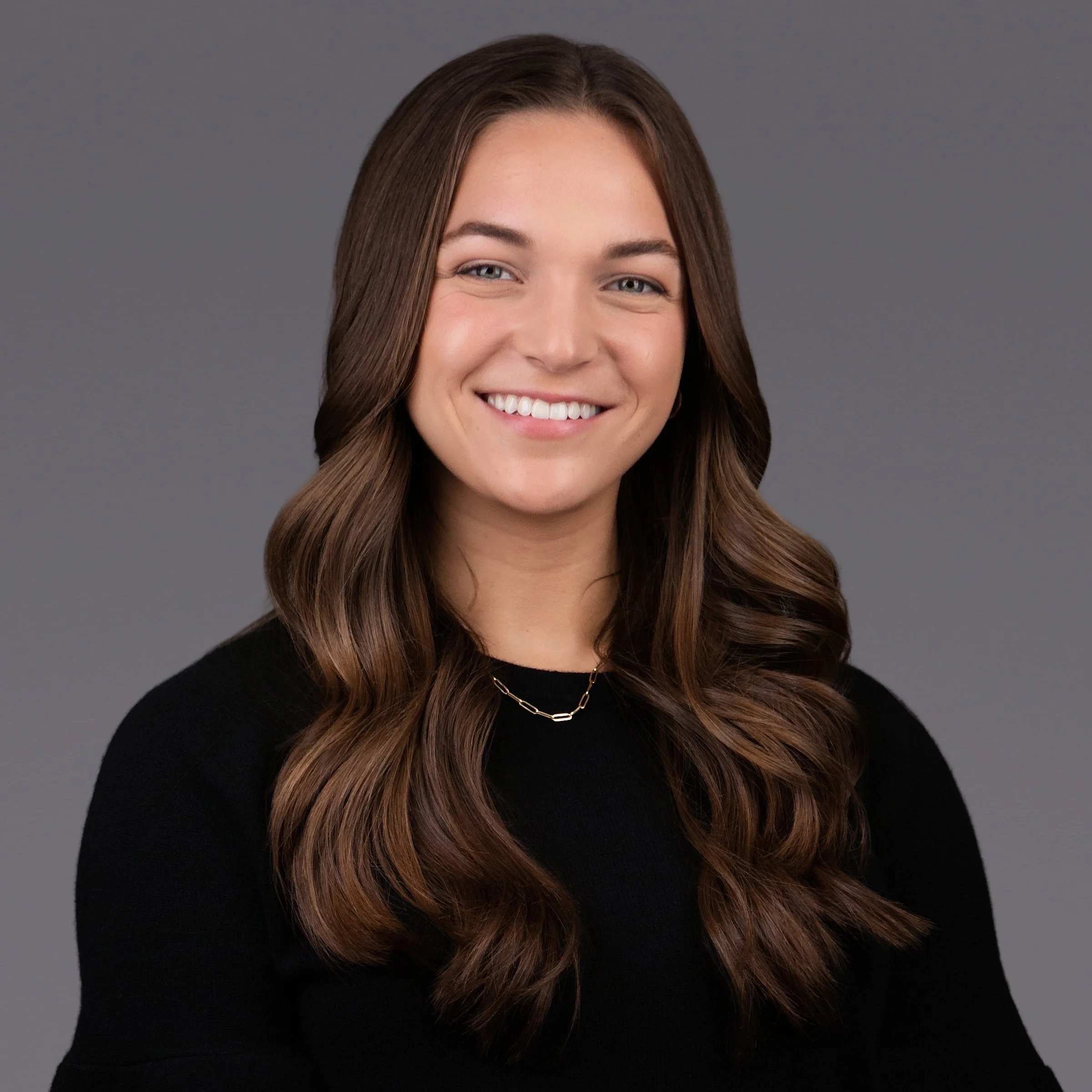 Professional corporate headshot of a smiling businesswoman with long brown hair, wearing a black top against a gray studio background, photographed by SoFlo Headshots in Miami, Florida.