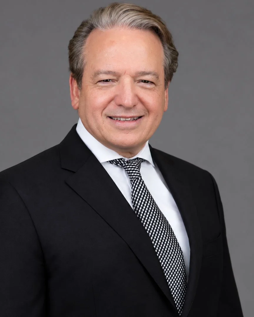 Professional corporate headshot of a smiling gray-haired businessman wearing a black suit, white shirt, and patterned tie against a neutral gray studio background.