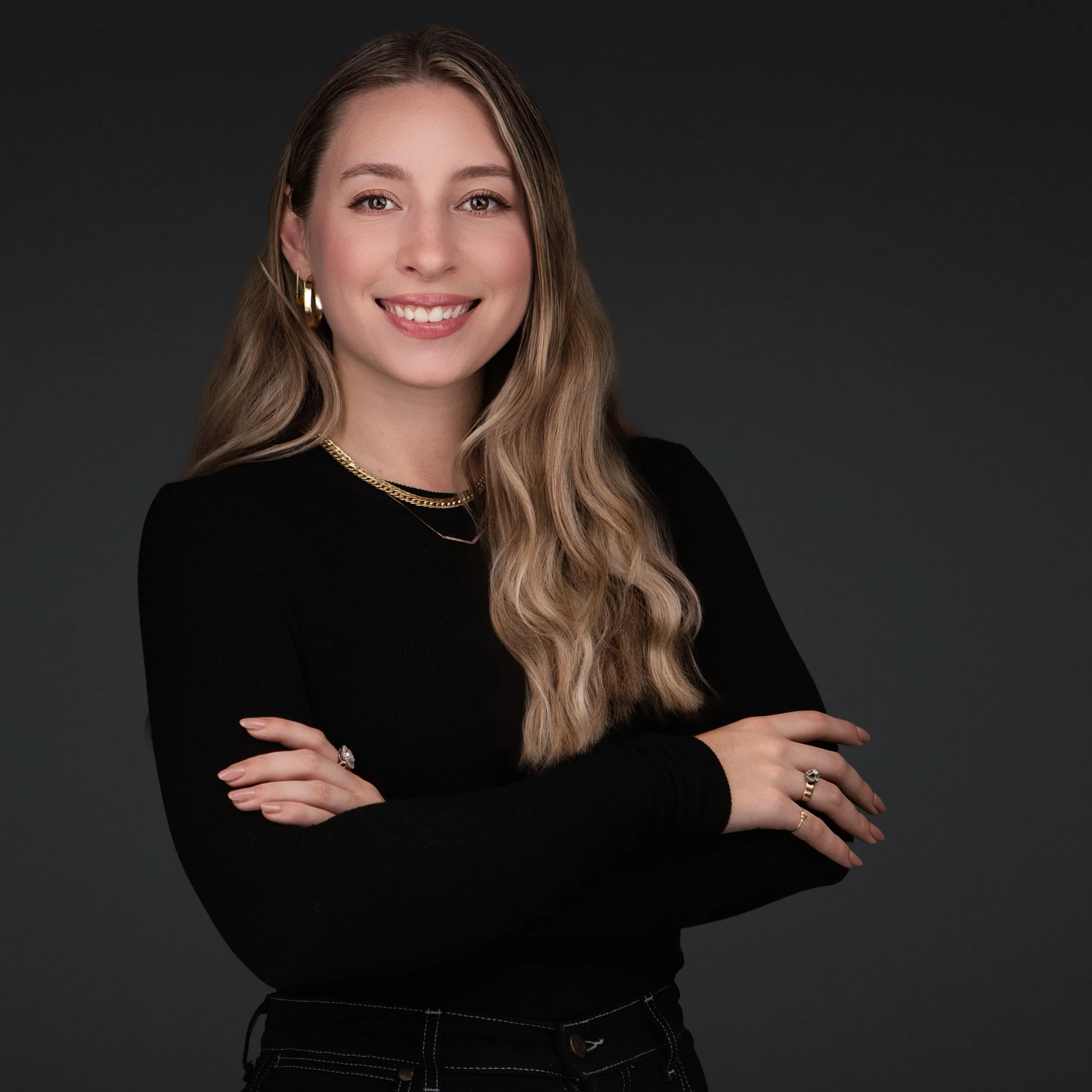 Professional female business headshot featuring long blonde hair, black attire, and a friendly smile against a dark background.