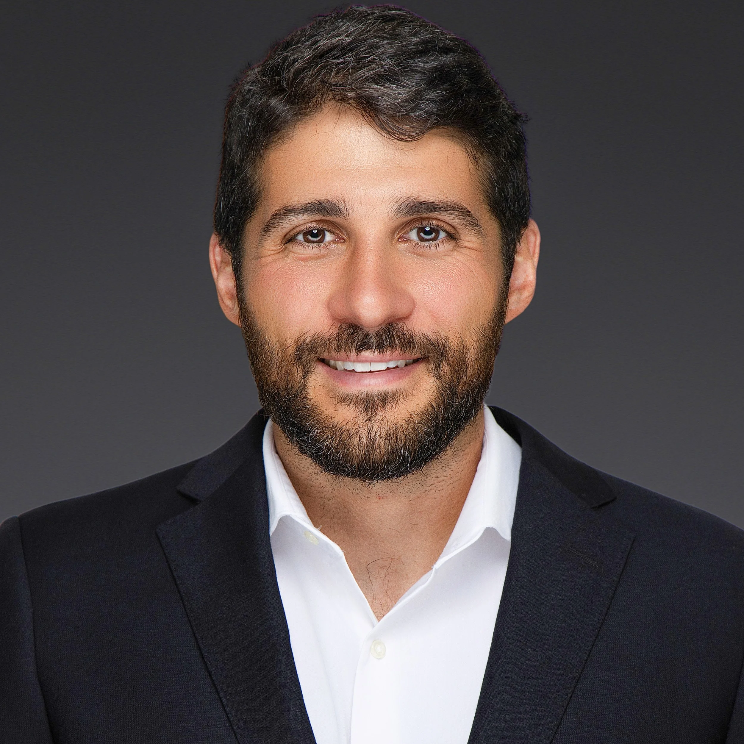 Professional corporate headshot of a smiling male executive with dark hair and a beard, wearing a black suit jacket and white dress shirt against a dark gray studio background, photographed by SoFlo Headshots in Miami, Florida.