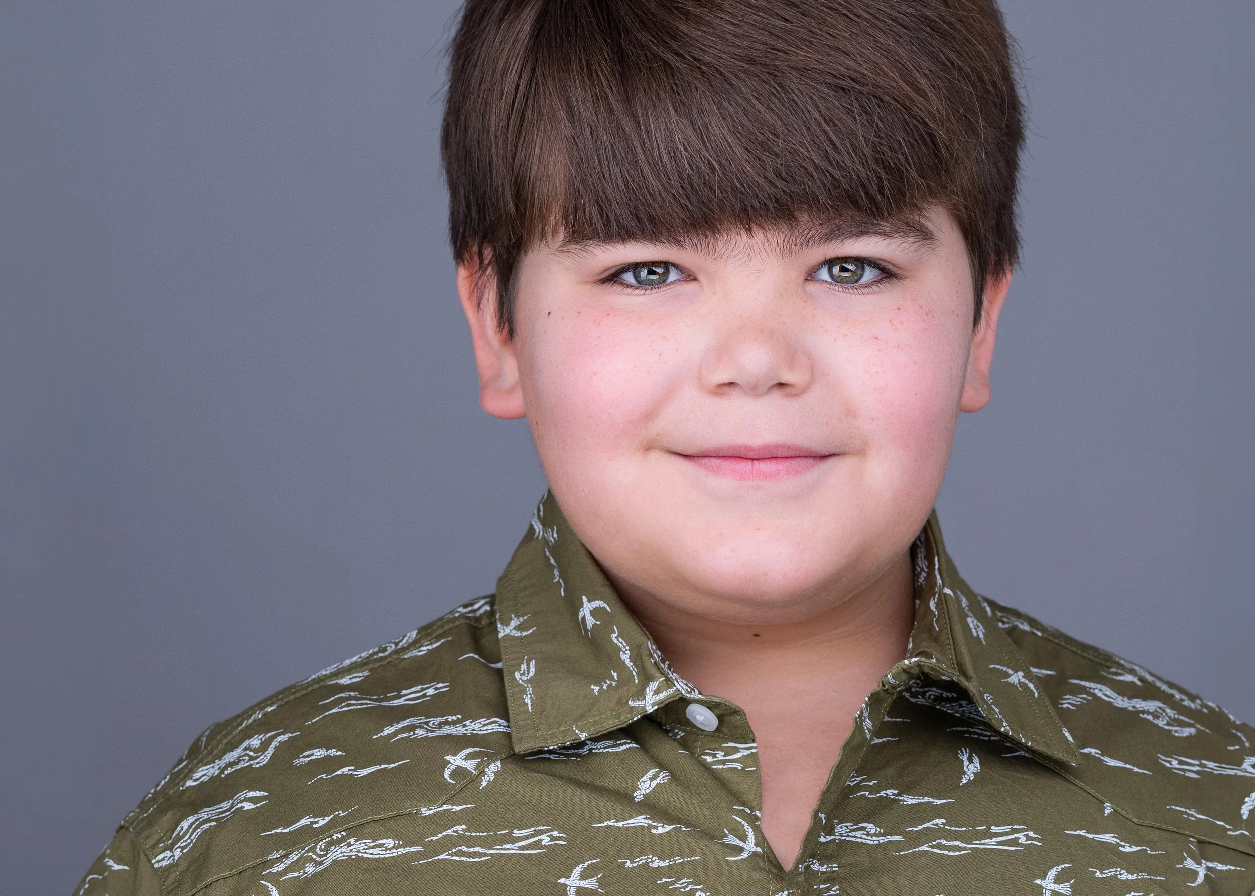 Young male actor headshot featuring a natural expression, patterned green shirt, and gray studio background.