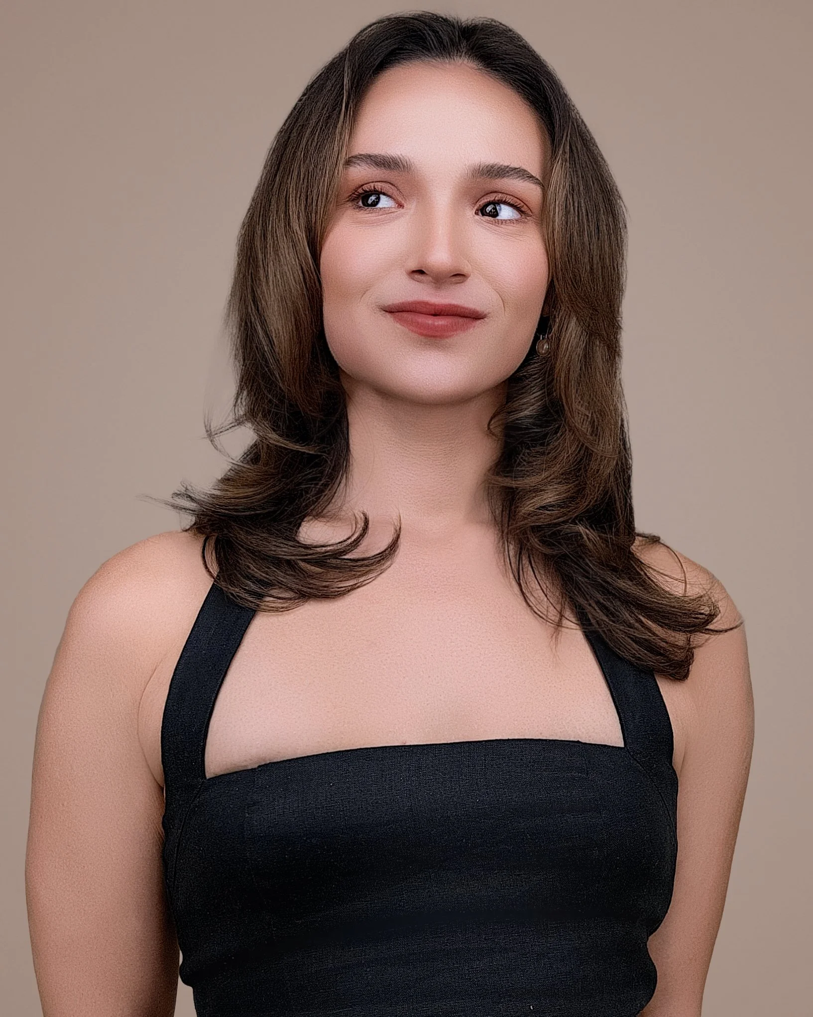 Female actor portfolio headshot featuring shoulder-length brown hair, a black top, and a warm neutral studio background.