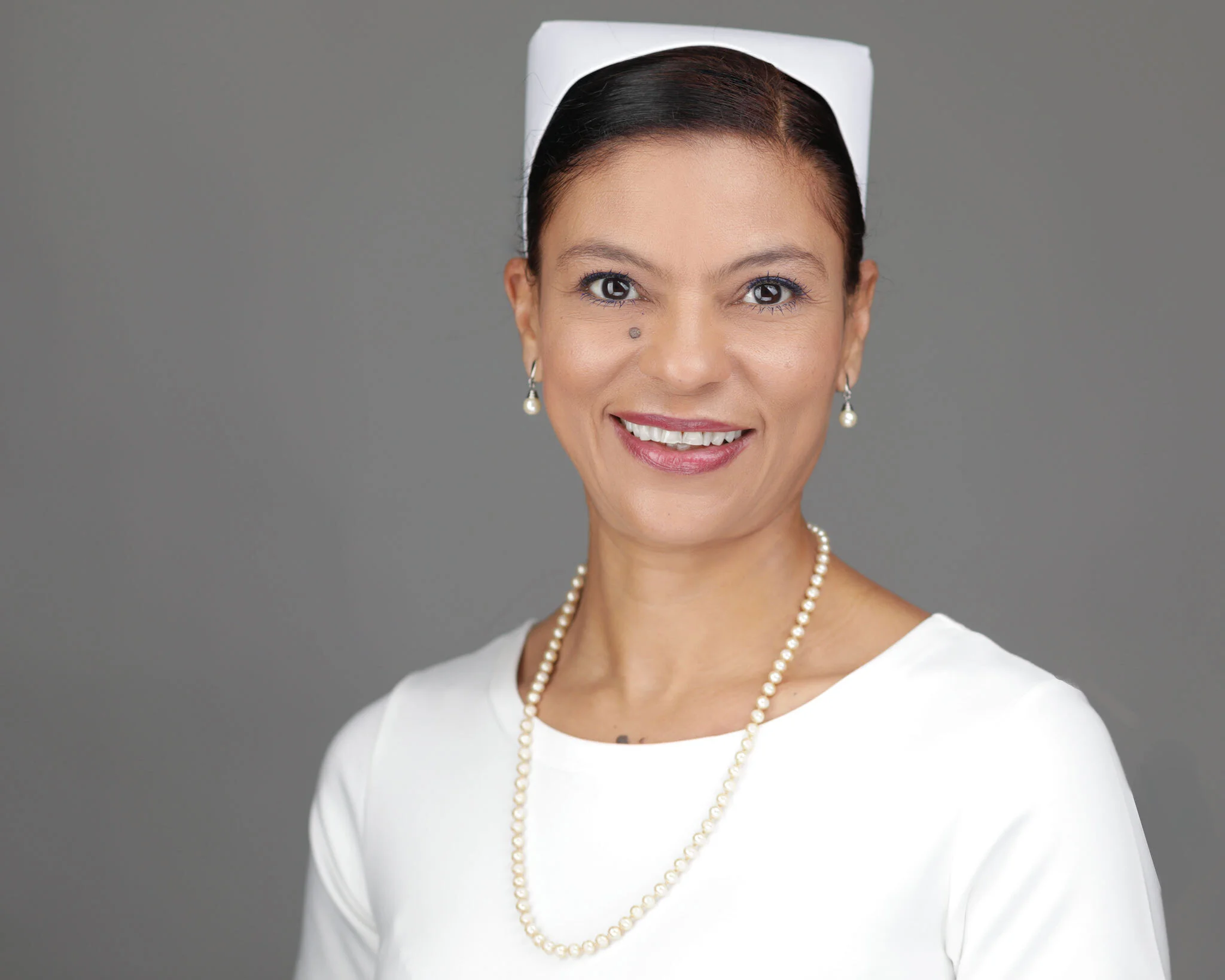 Professional woman's headshot featuring a white outfit, pearl necklace, and warm smile against a gray background.