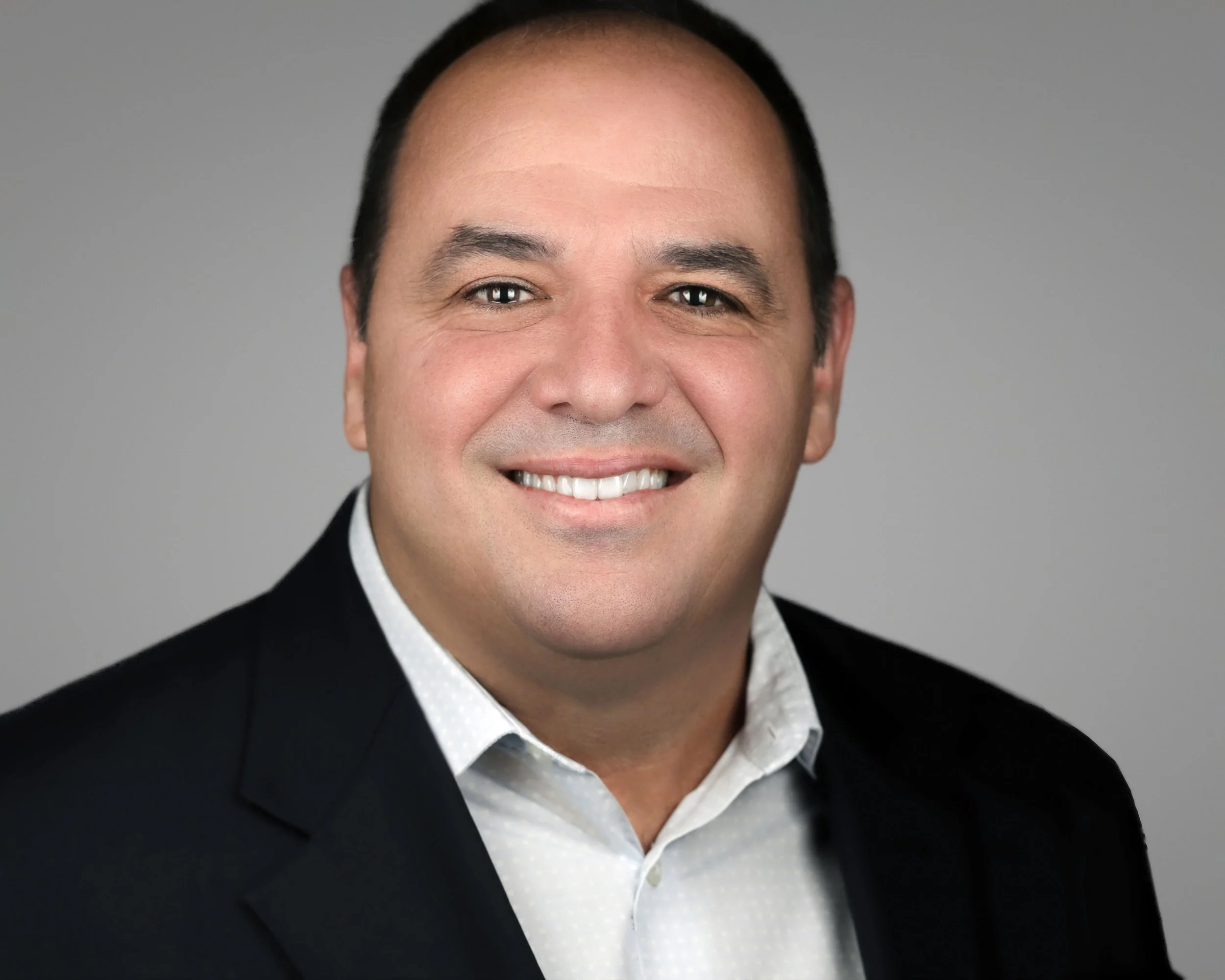 Professional male business headshot featuring a friendly smile, dark suit jacket, and gray studio background.