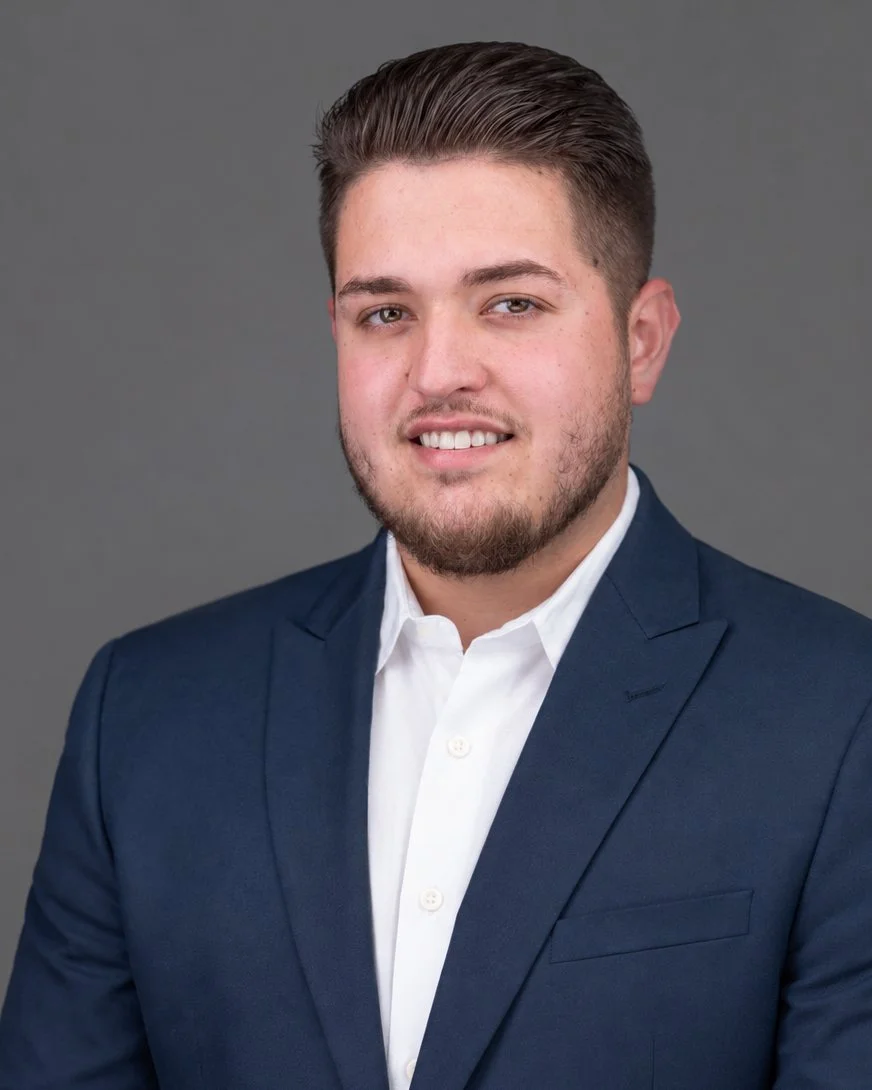 Professional corporate headshot of a male business executive wearing a navy blue suit and white shirt against a gray studio background, photographed by SoFlo-HeadShots in Miami, Florida.