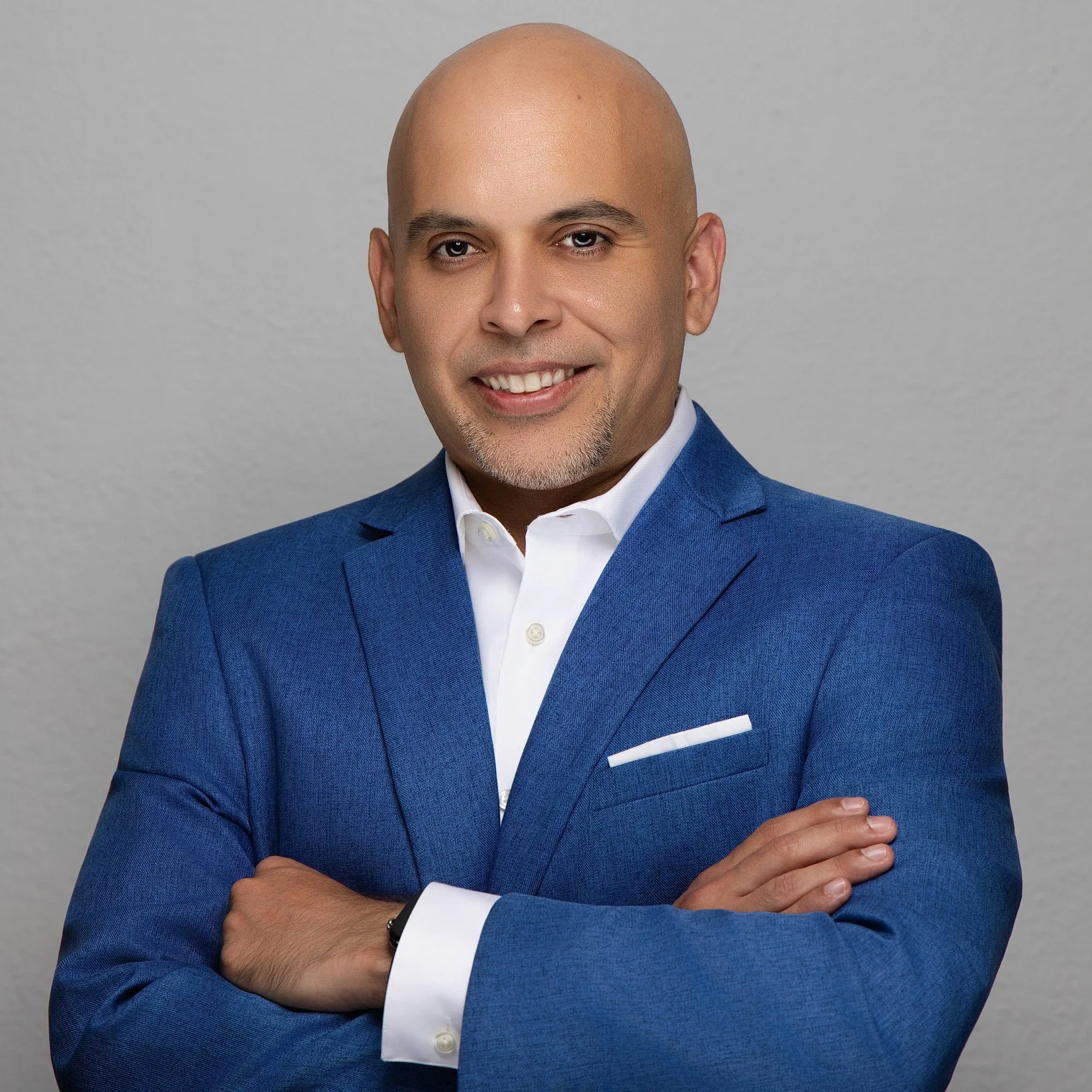 Professional corporate headshot of a smiling male business executive wearing a blue suit and white dress shirt, standing with arms crossed against a light gray background, photographed by SoFlo Headshots in Miami, Florida.