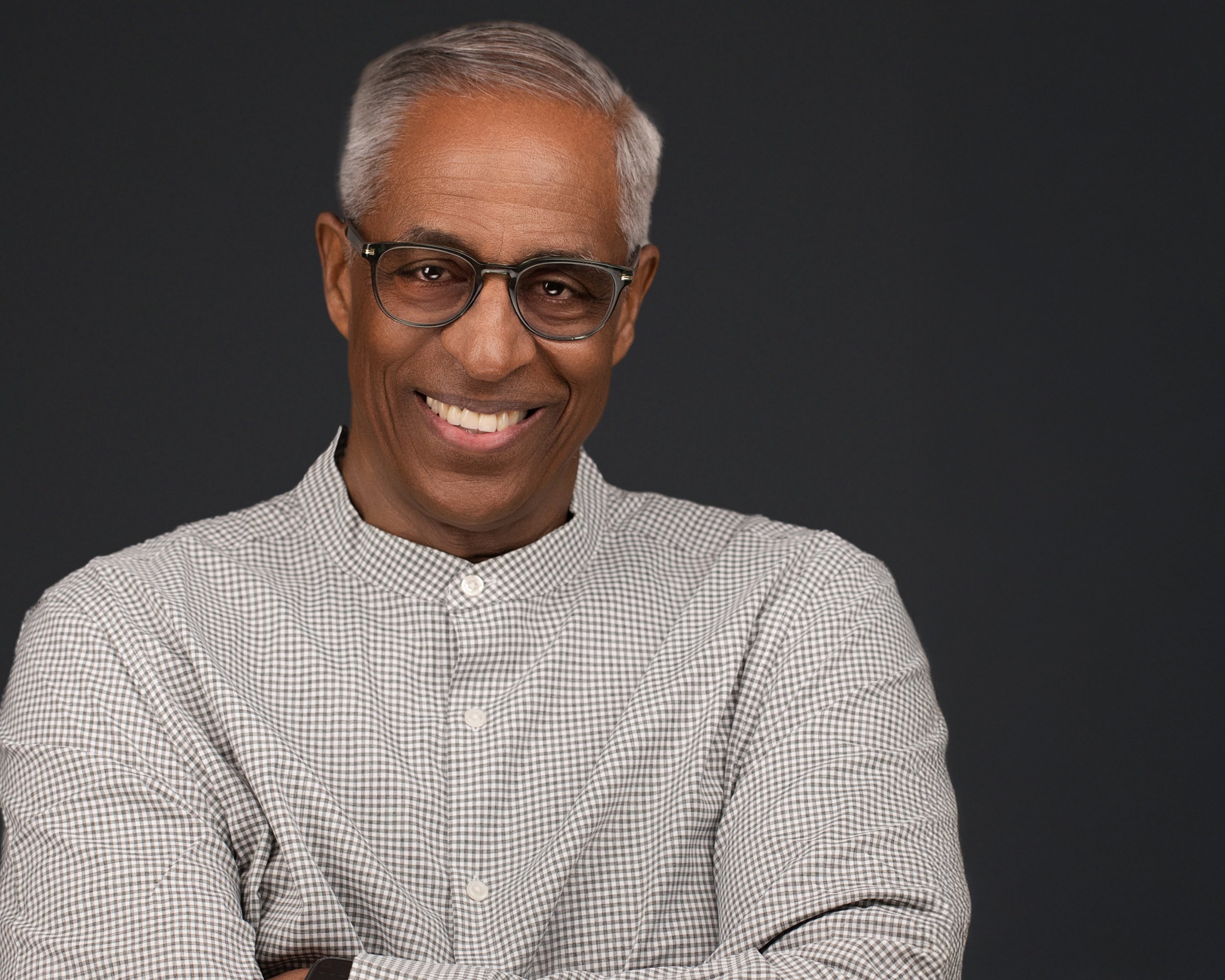 Professional headshot of a smiling older man wearing glasses and a light patterned shirt against a dark studio backdrop.