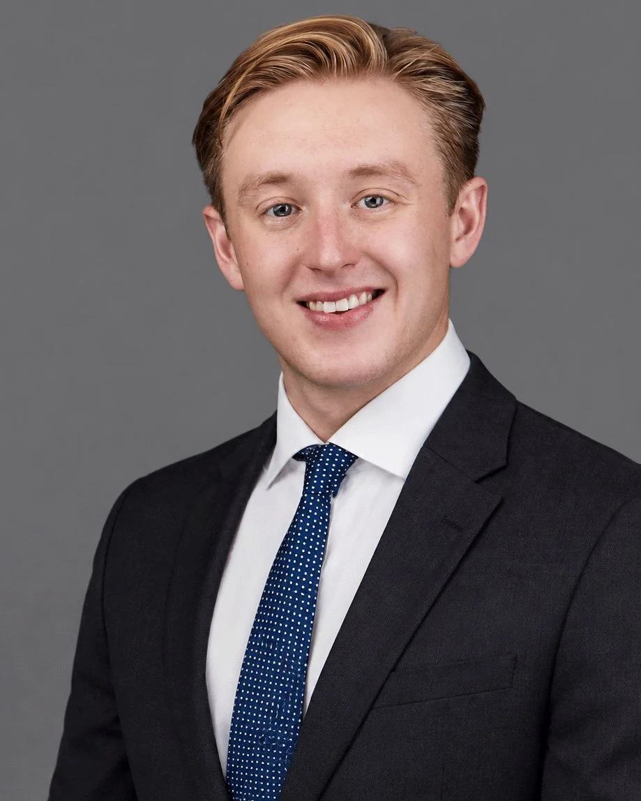 Professional corporate headshot of a smiling red-haired businessman wearing a dark suit, white shirt, and blue patterned tie against a gray studio background, photographed by SoFlo-HeadShots in Miami.