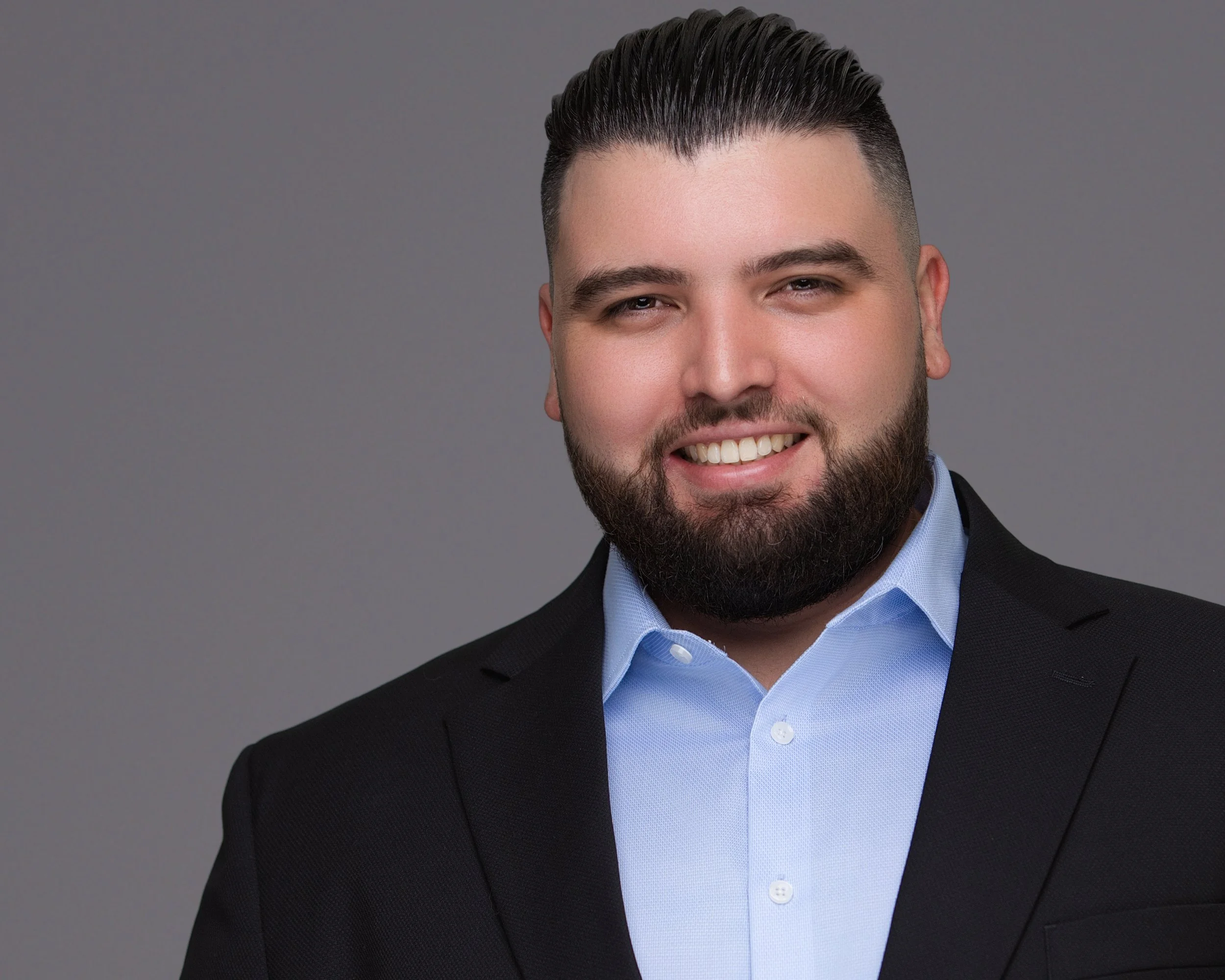 Professional male executive headshot featuring a dark suit, light blue shirt, and gray studio backdrop