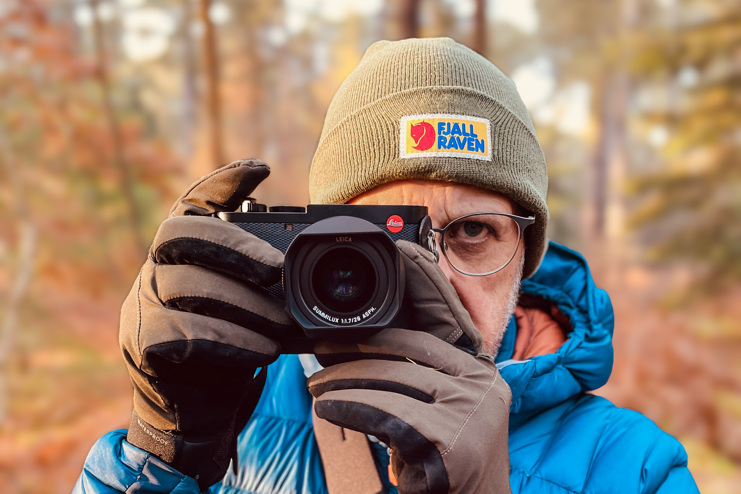 Person wearing glasses and a beanie, holding a Leica camera, in an outdoor setting.