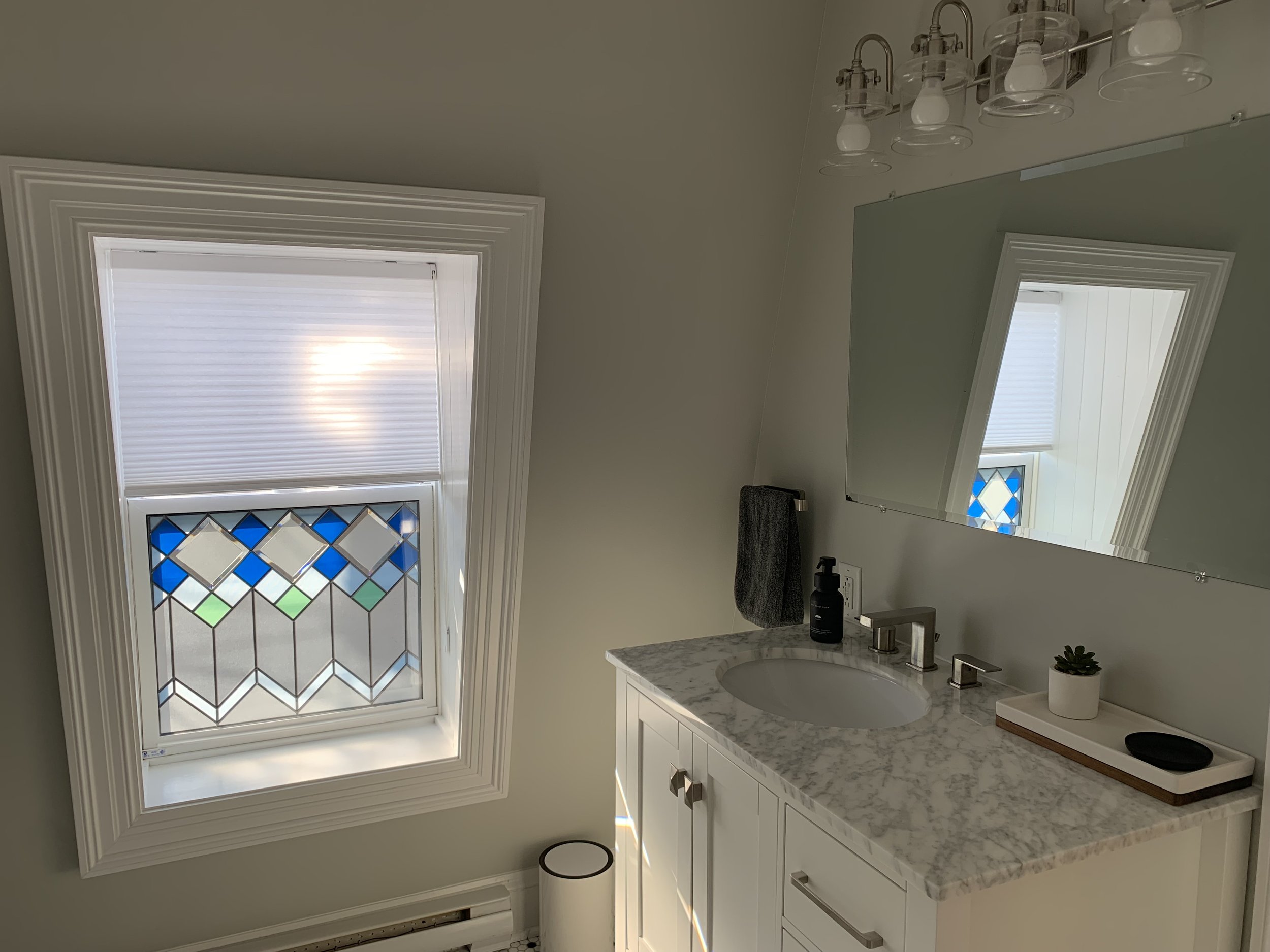 Bathroom with a white marble countertop, a sink with a modern faucet, a large mirror, a window with stained glass and white blinds, a black soap dispenser, a gray towel, a small potted plant, and a tray with items. The ceiling has a light fixture wit