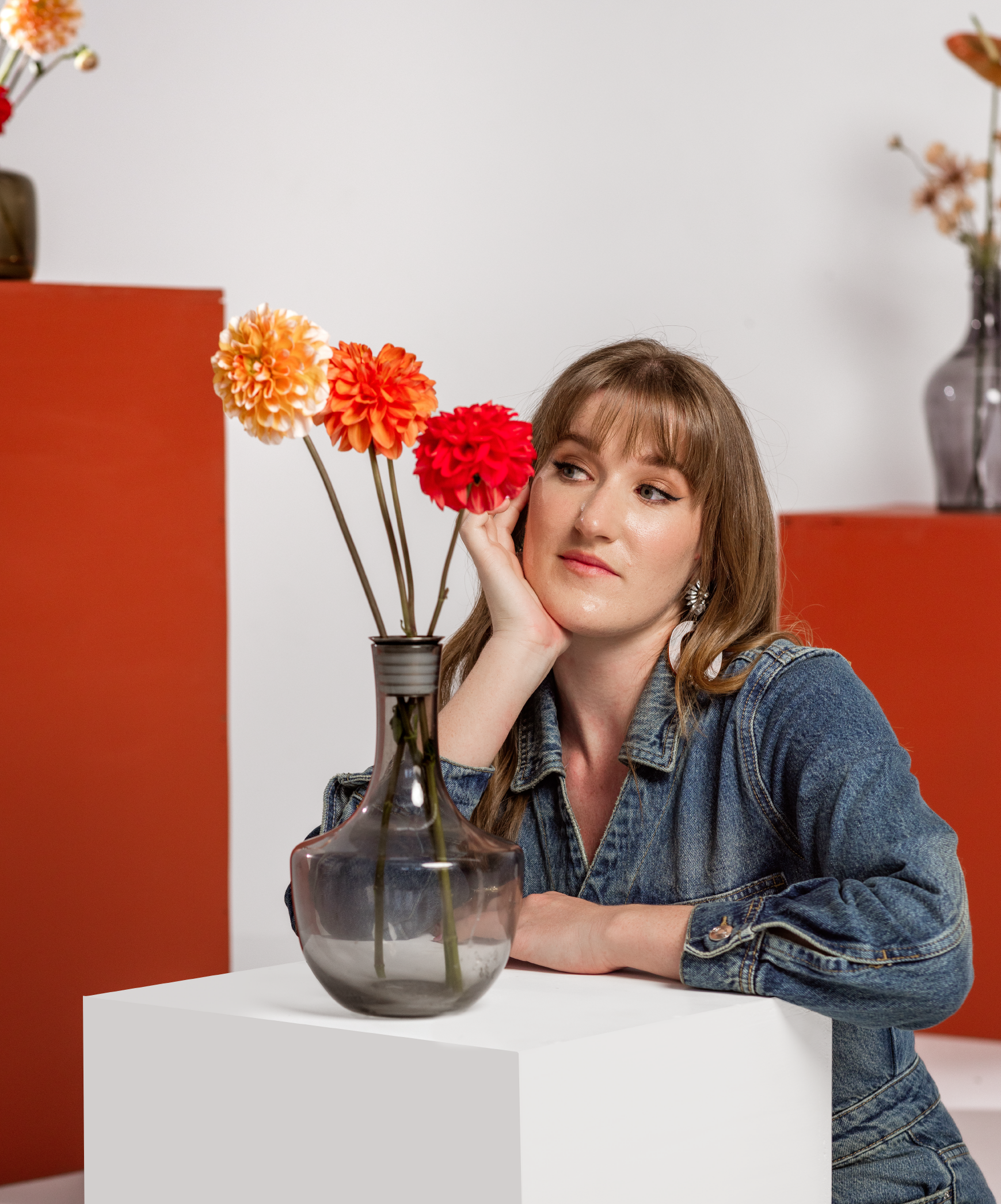 Person in denim jacket leaning on white pedestal with glass vase holding orange and red flowers, against a white background with red accents.