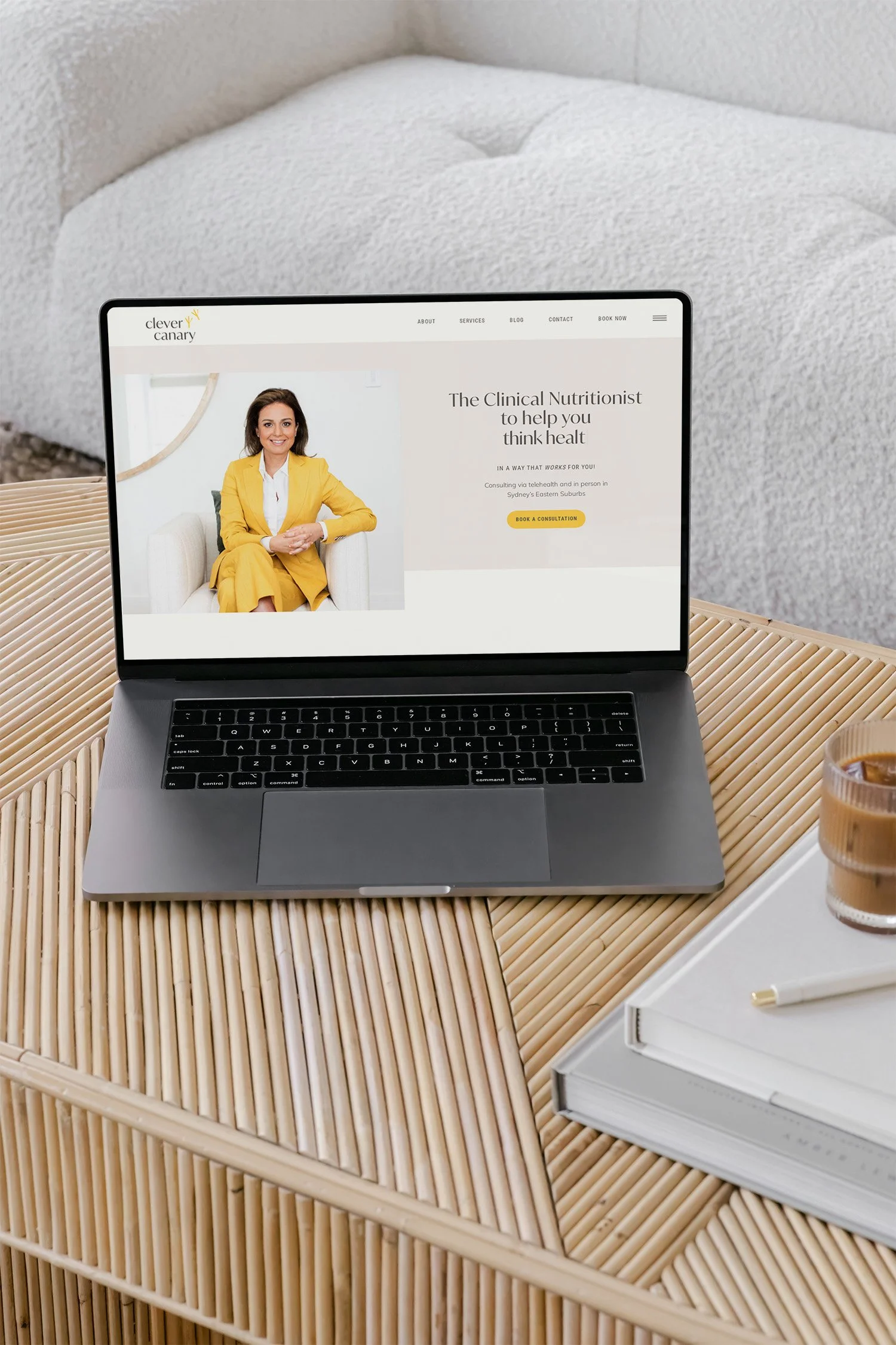 A laptop on a wooden table displaying a website for a clinical nutritionist, with a woman in a yellow blazer on the screen. Next to the laptop are a glass of iced coffee, a notebook, and a pen.