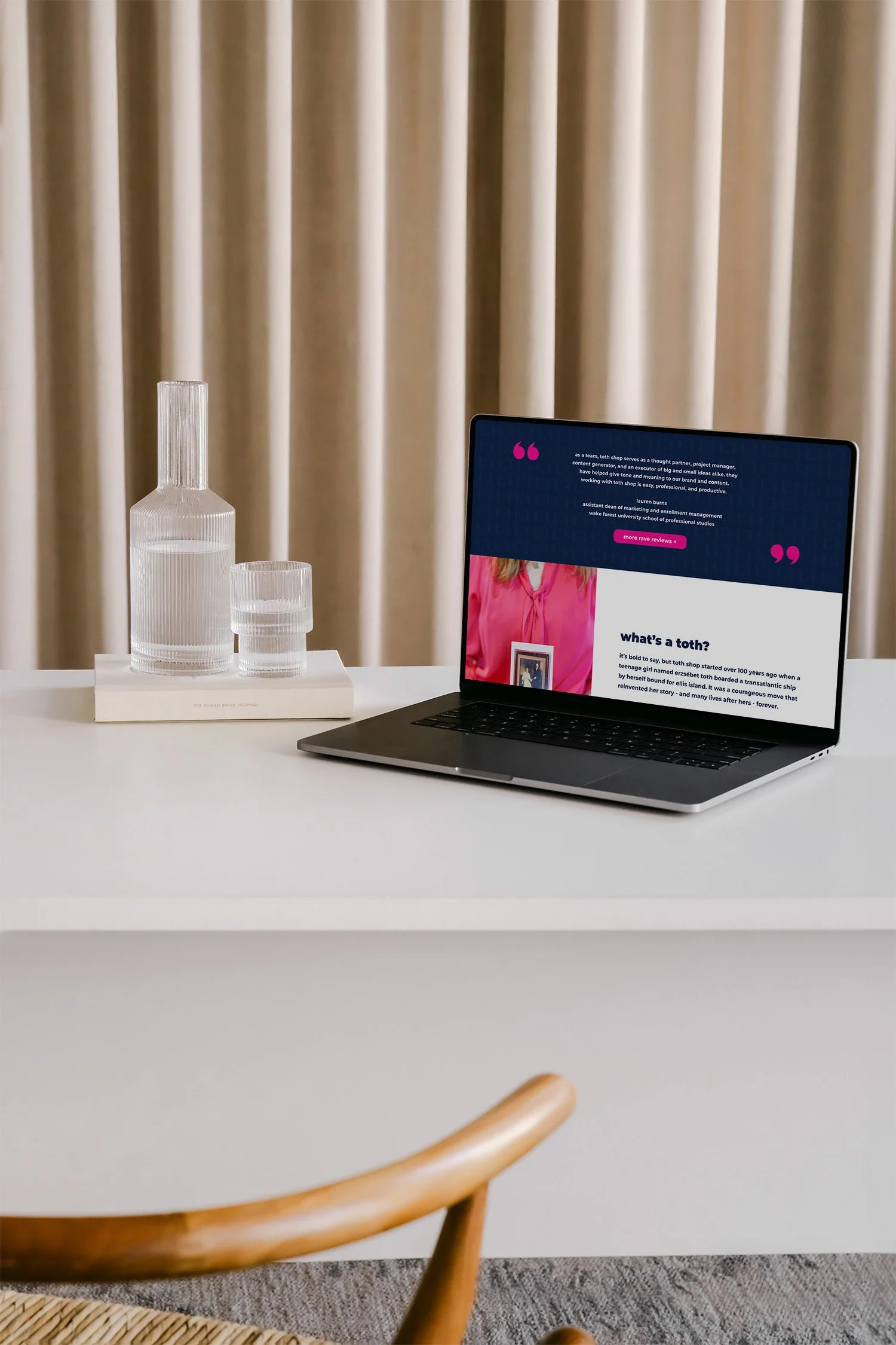 A laptop displaying a webpage sits on a white desk next to a glass water carafe and glass, with beige curtains in the background.