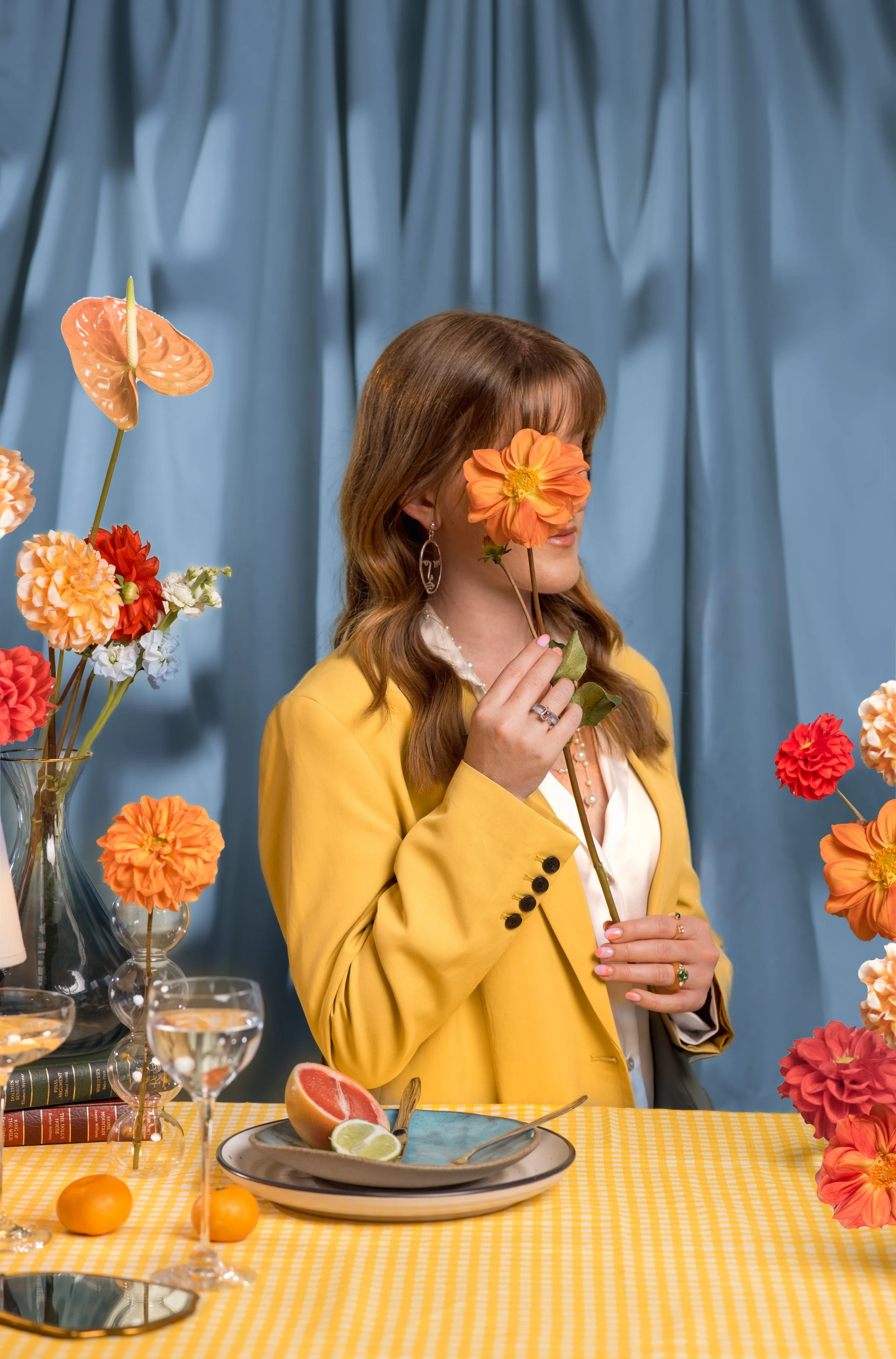 A woman with red hair, wearing a yellow blazer, holding a large orange flower in front of her face, seated at a table with colorful flowers, fruit, and glasses of water against a blue curtain background.
