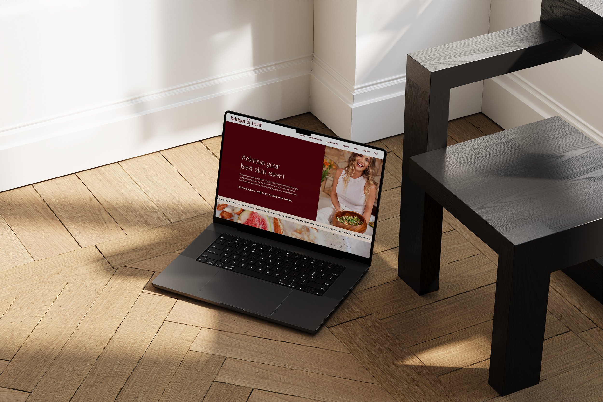 Laptop on a wooden floor displaying a skincare website with a smiling woman holding a salad near a black table.