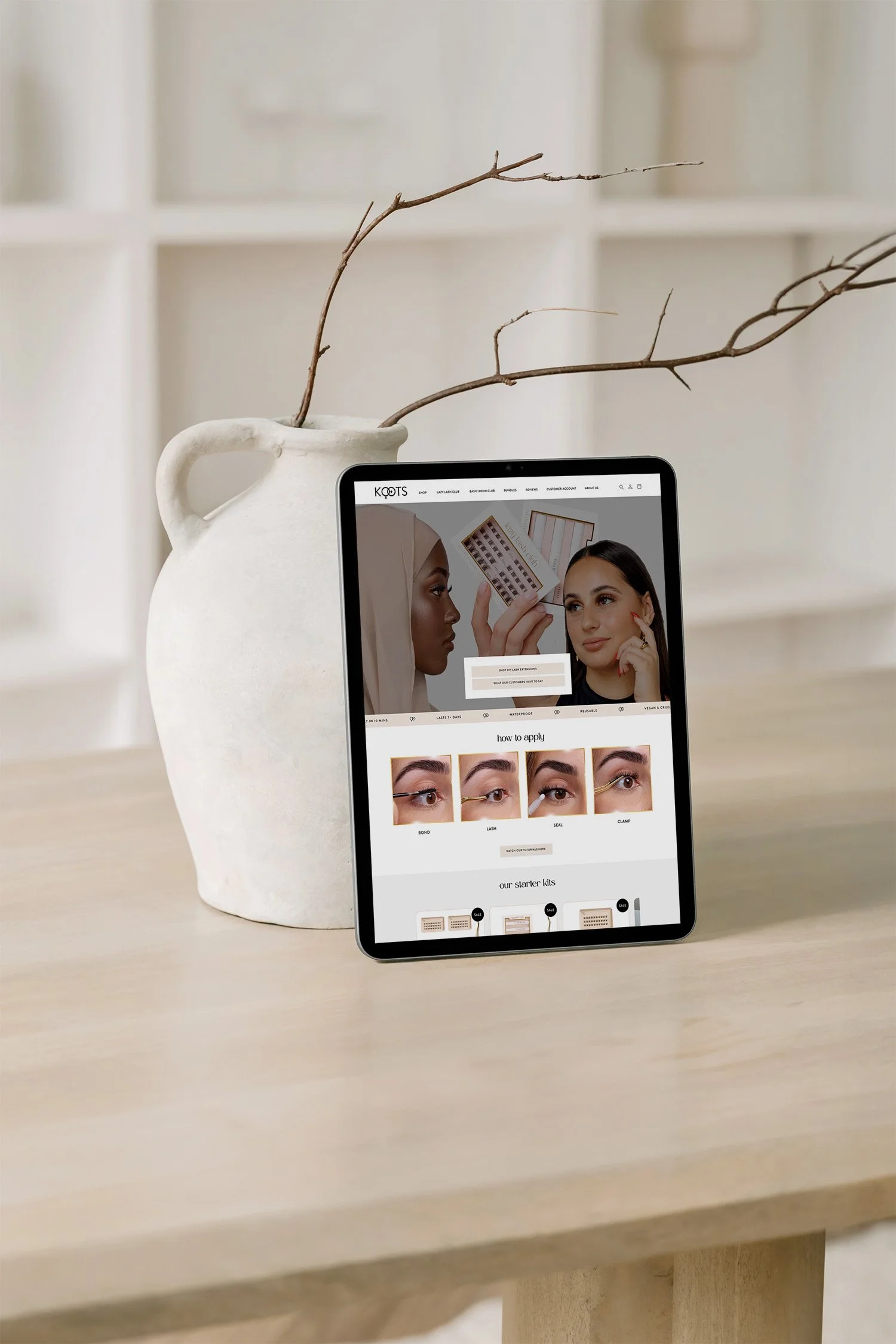 Tablet displaying a beauty website titled KOITS, showing images of women applying makeup, placed on a light wood table next to a white ceramic vase with dry branches inside.