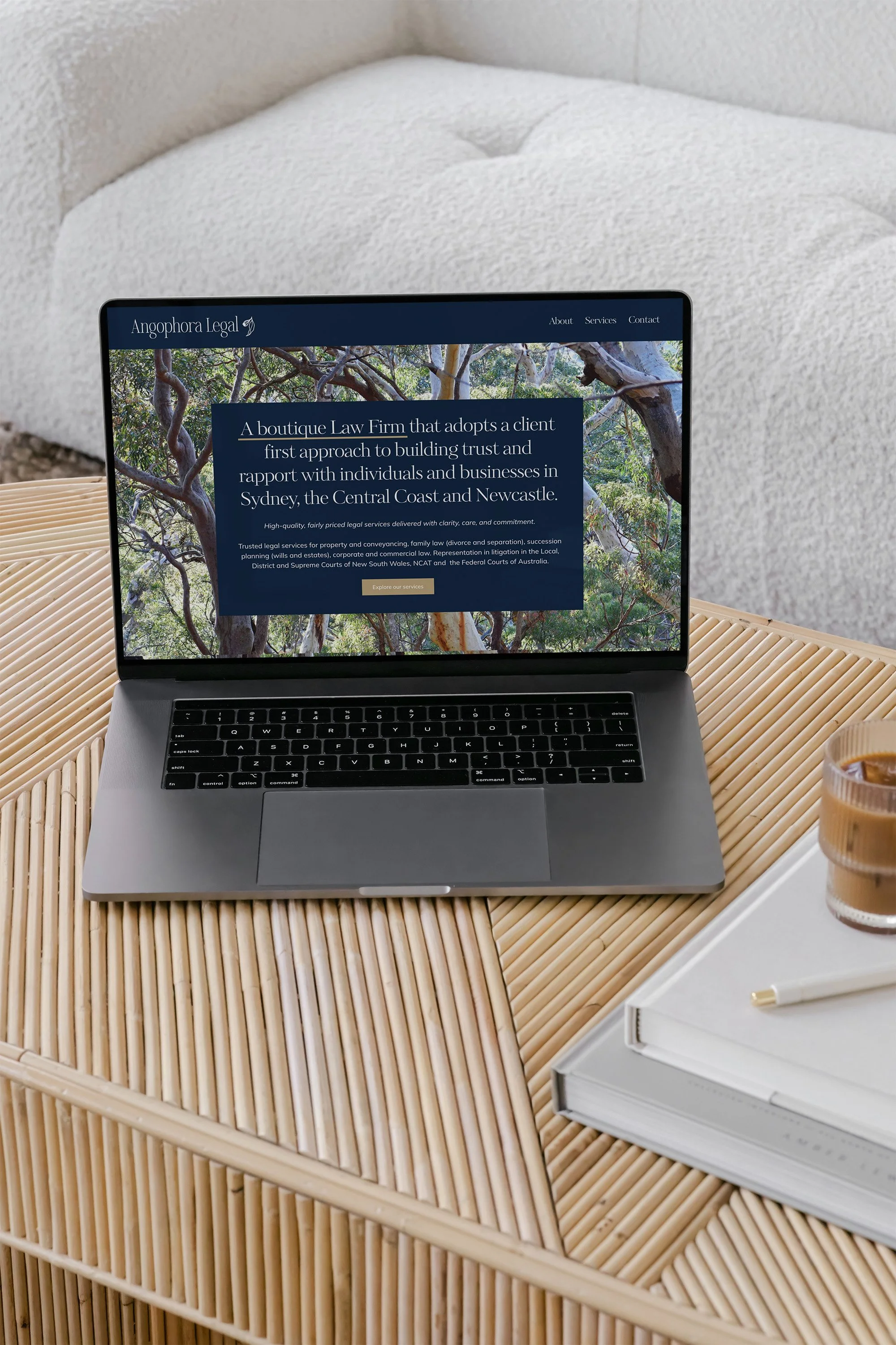 A laptop on a wooden desk displaying a legal firm's website, with a white couch and a glass of what appears to be coffee nearby.
