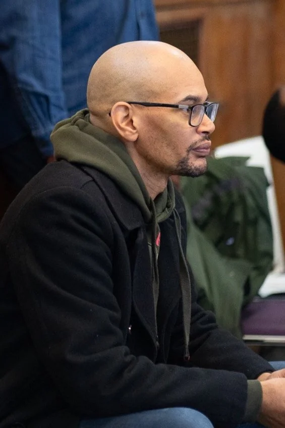Portrait of a person with short hair, wearing a patterned outfit and necklace, against a red background.