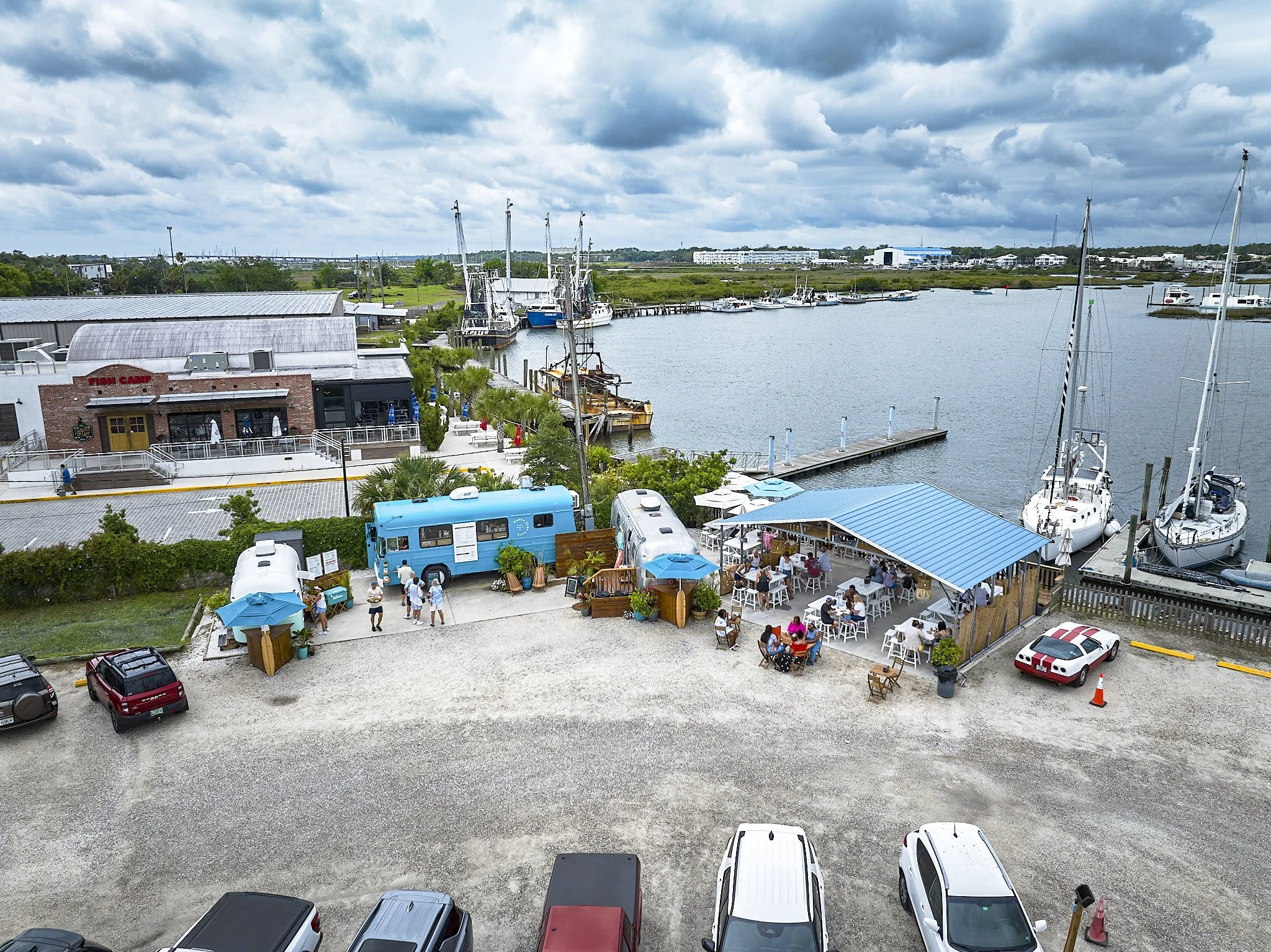 A waterfront scene with boats docked along a pier, a restaurant with outdoor seating, parked cars, and a cloudy sky overhead.