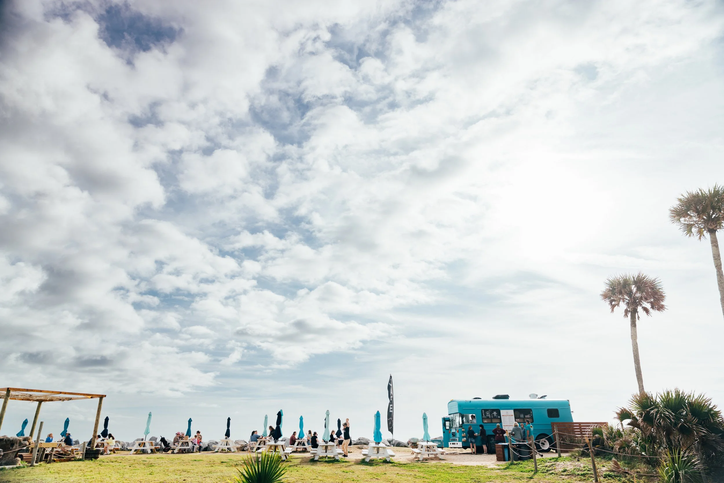 Beach scene with people sitting at picnic tables, blue umbrellas, palm trees, and a food truck under a partly cloudy sky.