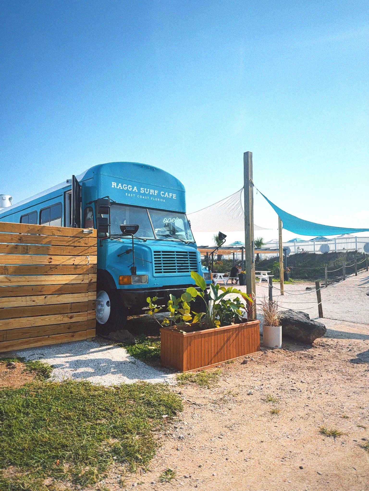 A bright blue food truck with the sign 'RAGGA SURF CAFE' parked behind a wooden fence on a sunny day at the beach. There are large white and blue shade sails above and a few people in the background. Potted plants and greenery are in the foreground.