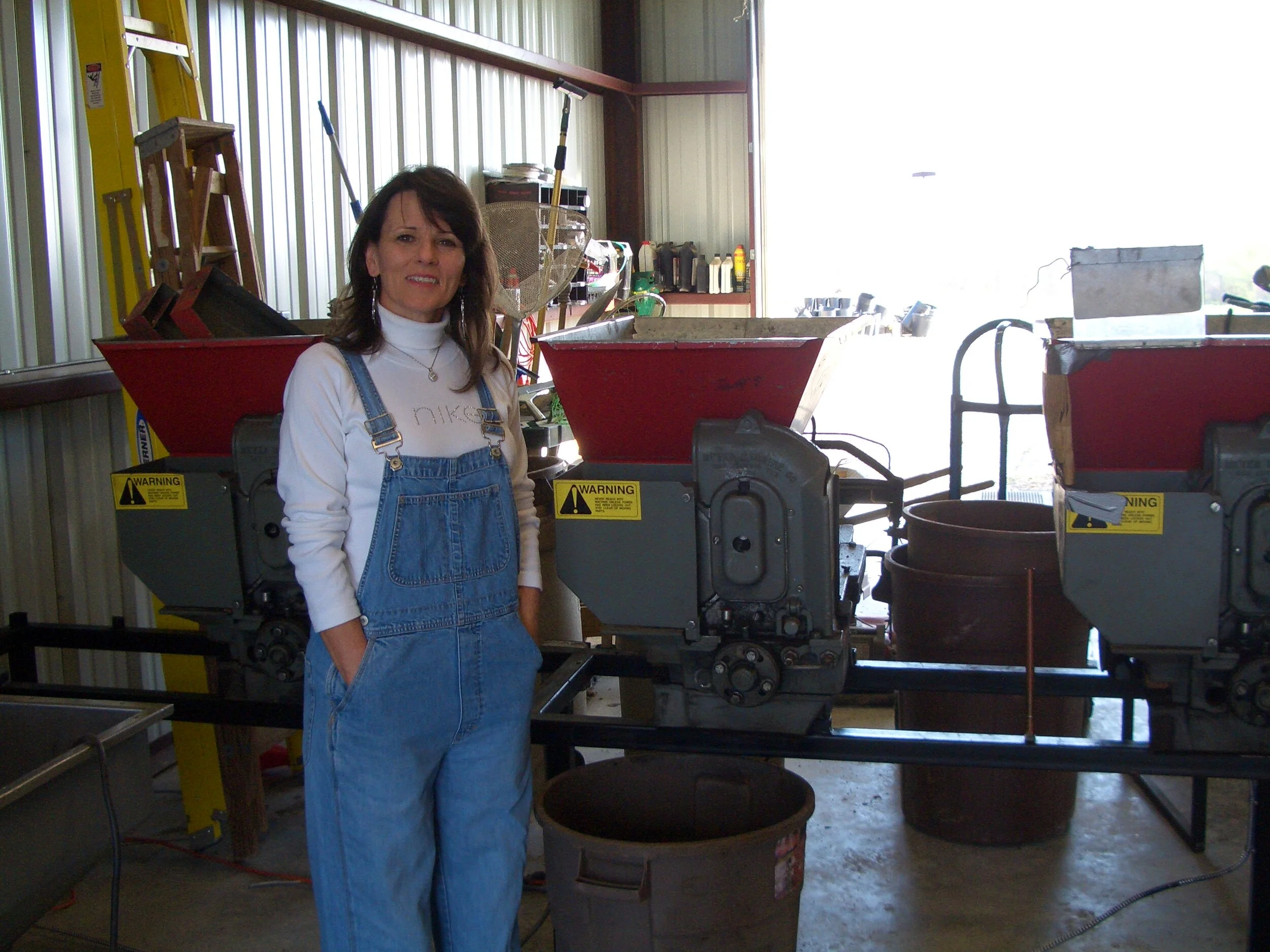 A woman in overalls standing inside a workshop with machinery and tools, some red containers, and a yellow ladder visible in the background.