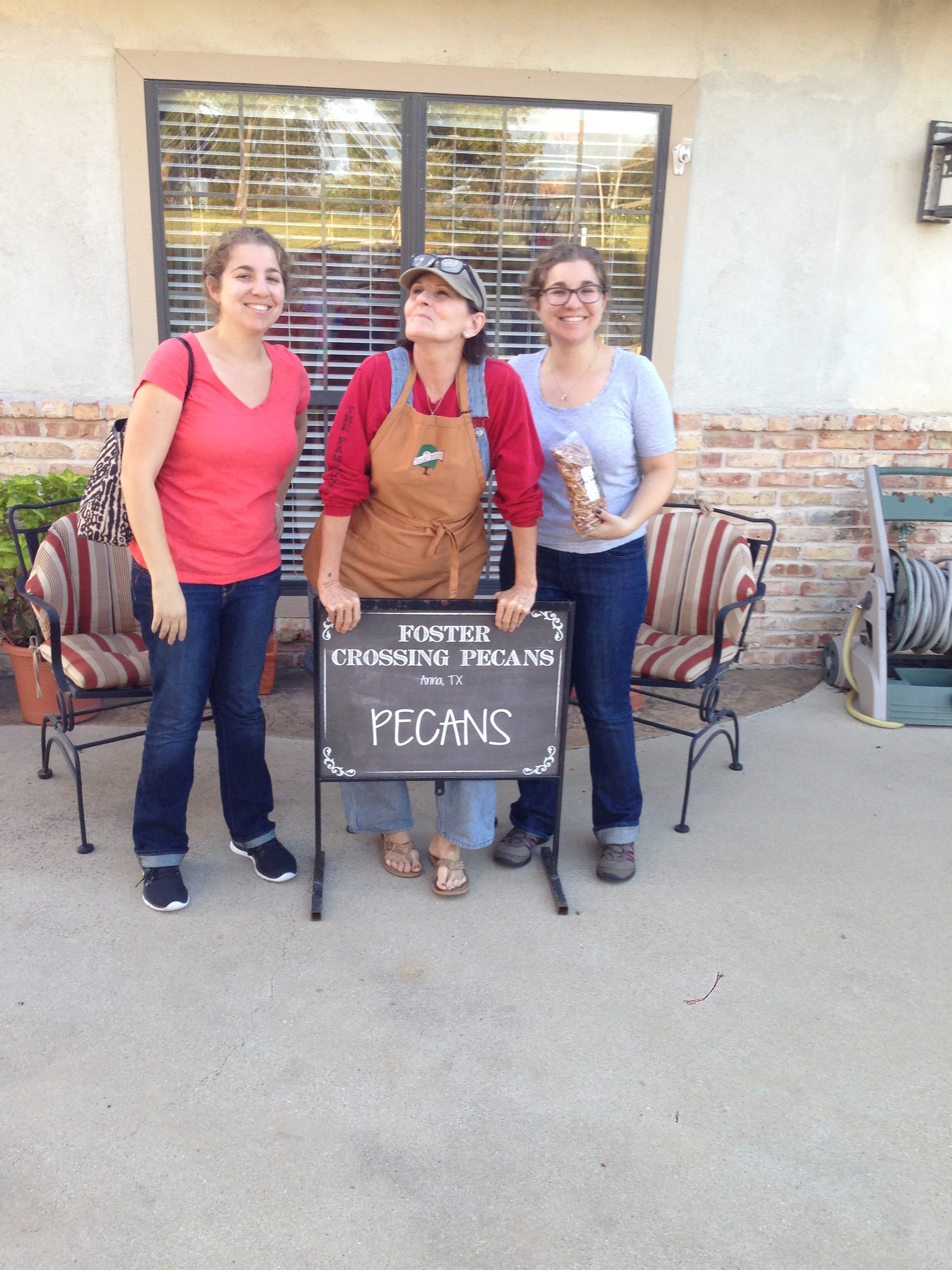 Three women standing outside, smiling, with a chalkboard sign that reads "Foster Crossing Pecans, Anna, TX, Pecans." One woman in the middle is wearing an apron and holding a bag of pecans.
