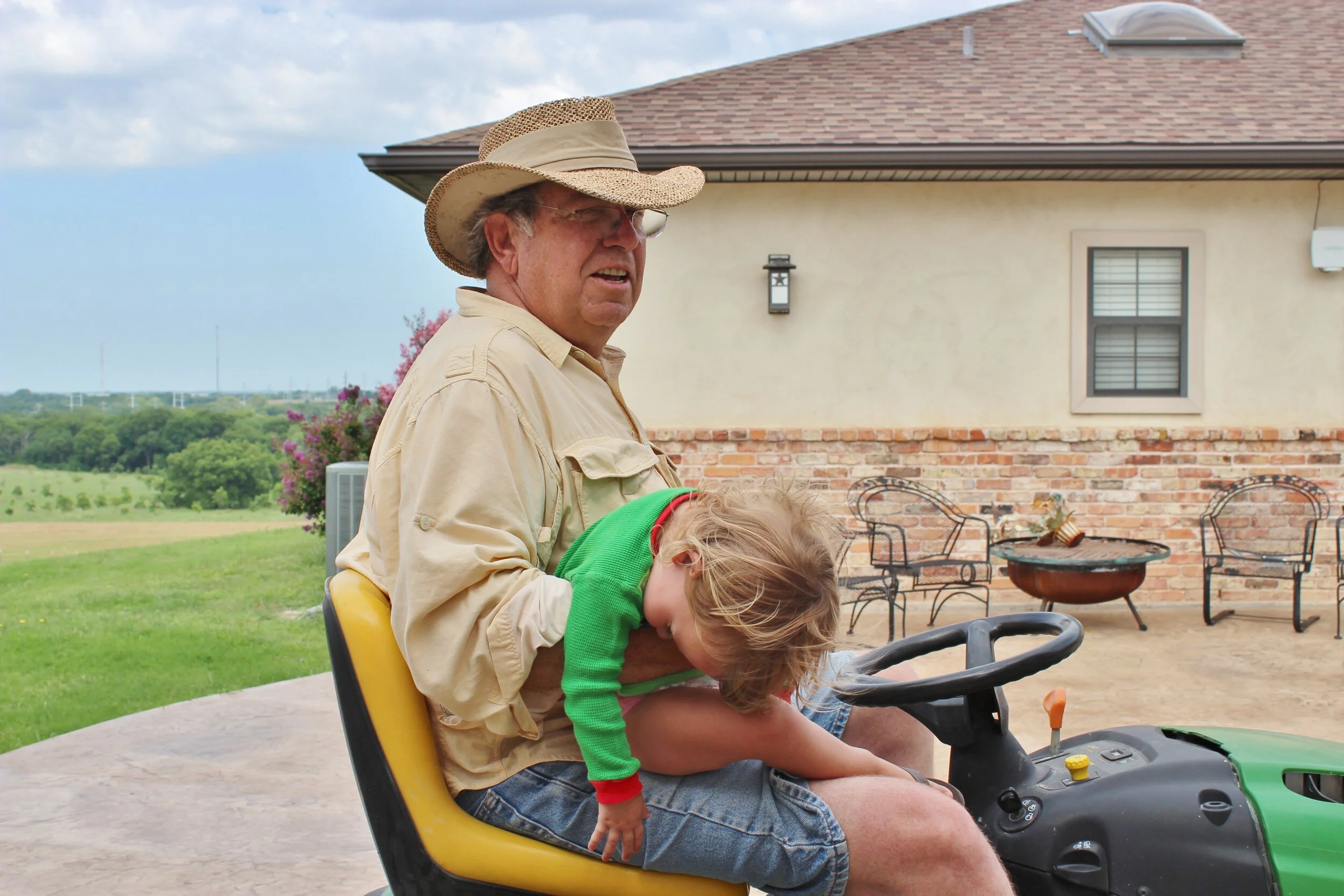 An elderly man wearing a straw hat and beige shirt sitting on a lawn tractor with a young boy in a green sweater and denim shorts resting on his lap, outdoors in front of a house with a brick and stucco wall and a patio with chairs and a fire ring.