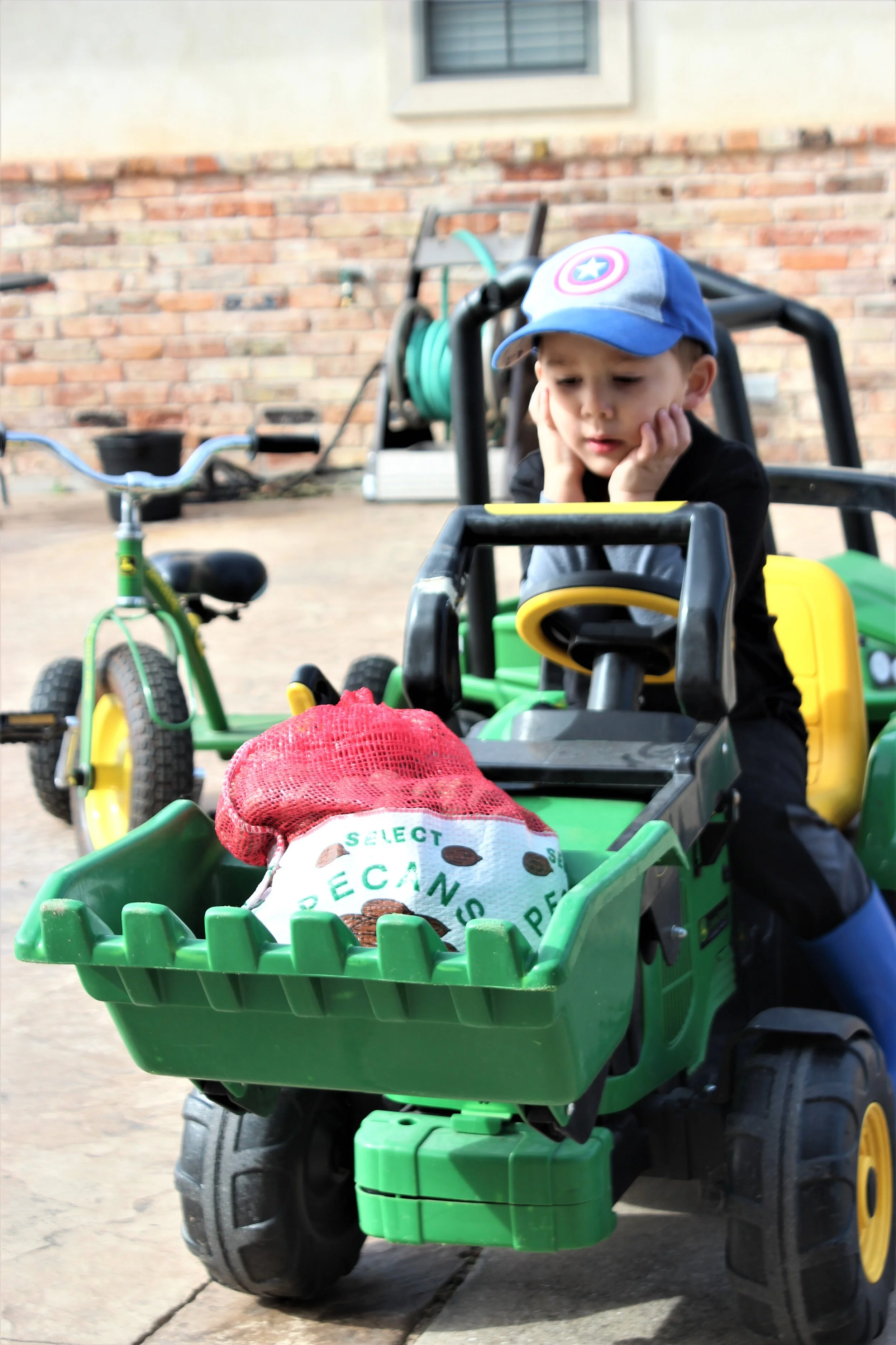 A young boy with a Captain America hat sits in a green toy tractor, looking thoughtful with his chin resting on his hands. There are other toys, including a tricycle, and gardening tools in the background. A red mesh bag containing pecans is in the f