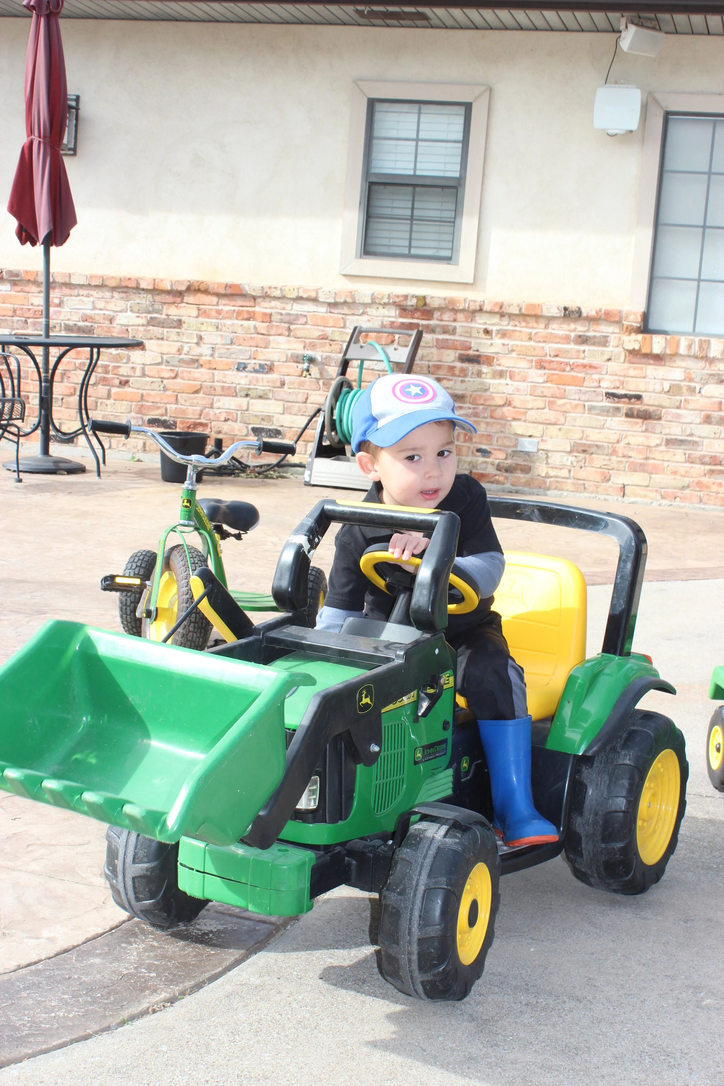Young boy wearing a Captain America cap and blue boots sitting in a green and yellow toy tractor in an outdoor play area.
