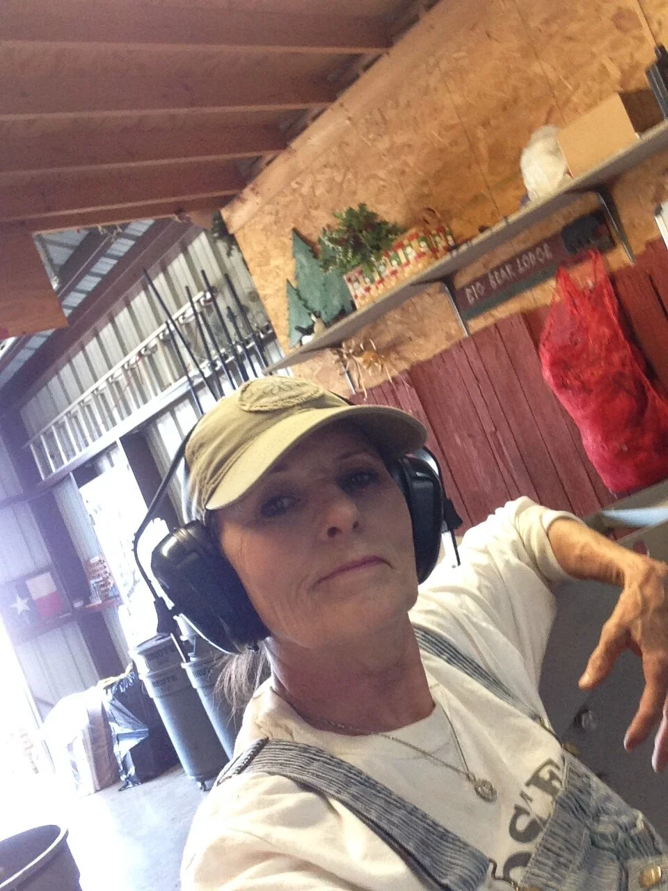 A woman wearing a beige baseball cap, large headphones, and overalls taking a selfie in a rustic indoor setting with wooden walls and ceiling, decorated with plants and Christmas items.