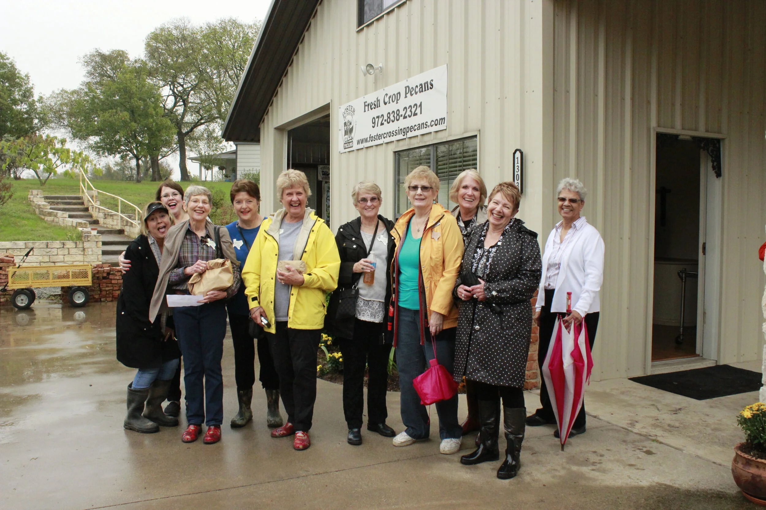 Group of women smiling and posing in front of a building with a sign that reads 'Fresh Crop Pecans'. It is a rainy day and some women are holding umbrellas.