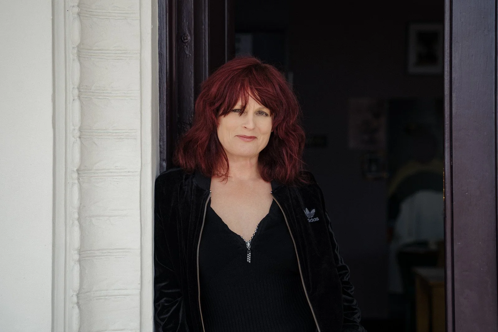 A woman with red hair standing in a doorway, wearing a black Adidas jacket and black top.