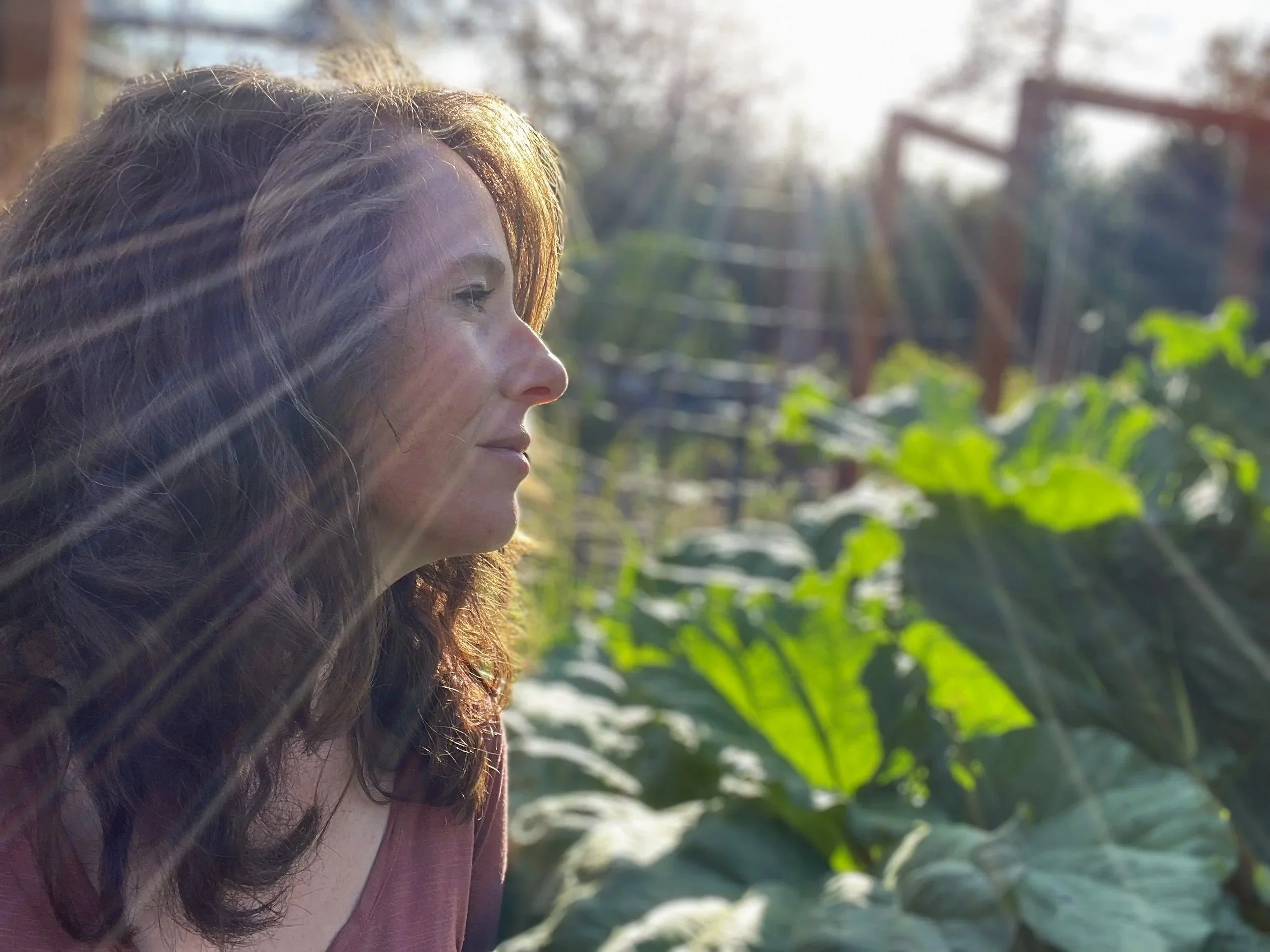 Esther Felts, Owner of Esther's Eden smiling with eyes closed, sitting in a garden of rhubarb leaves during late afternoon sunlight.