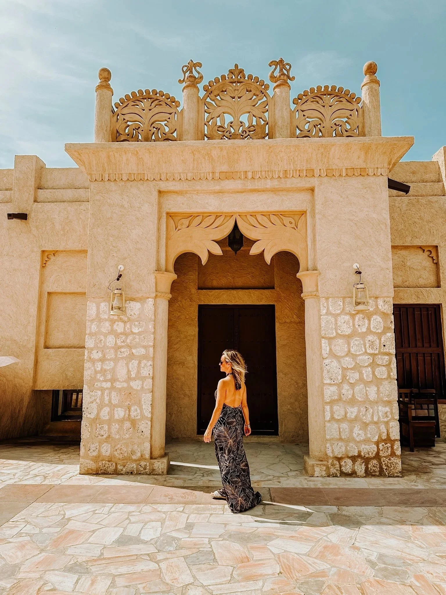 A stylish woman on vacation in a black and white patterned dress and sunglasses stands in front of an ornate beige stone building with intricate carvings and a large arched doorway in Dubai