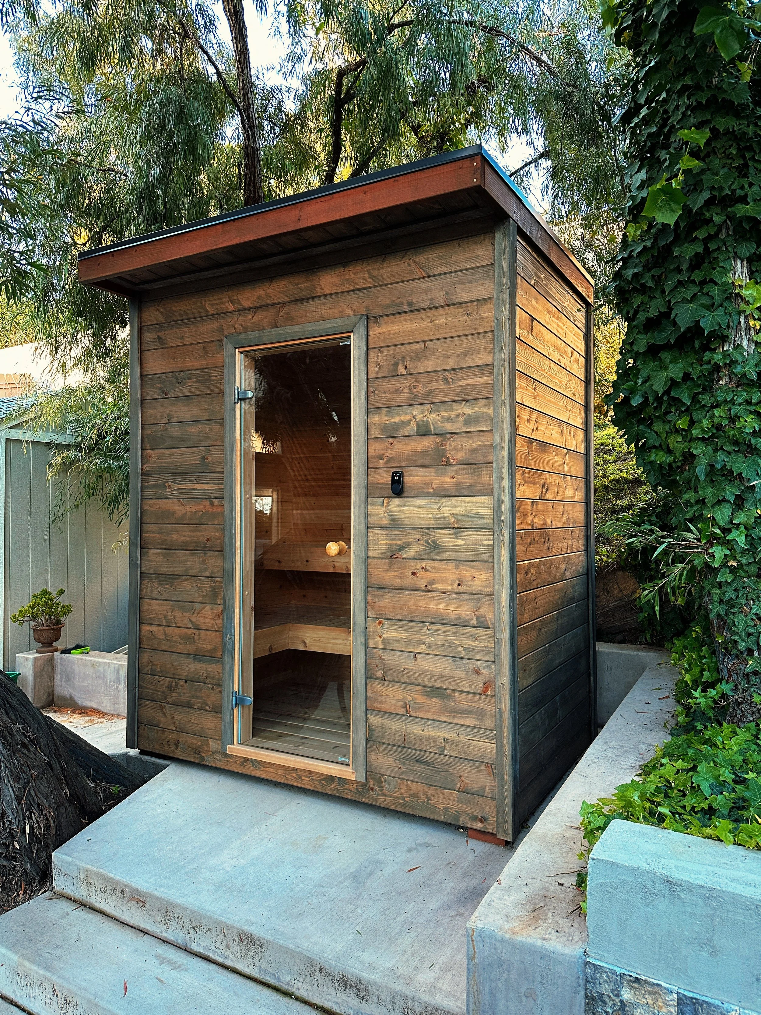 Small wooden outdoor sauna with glass door, located on concrete platform surrounded by greenery.