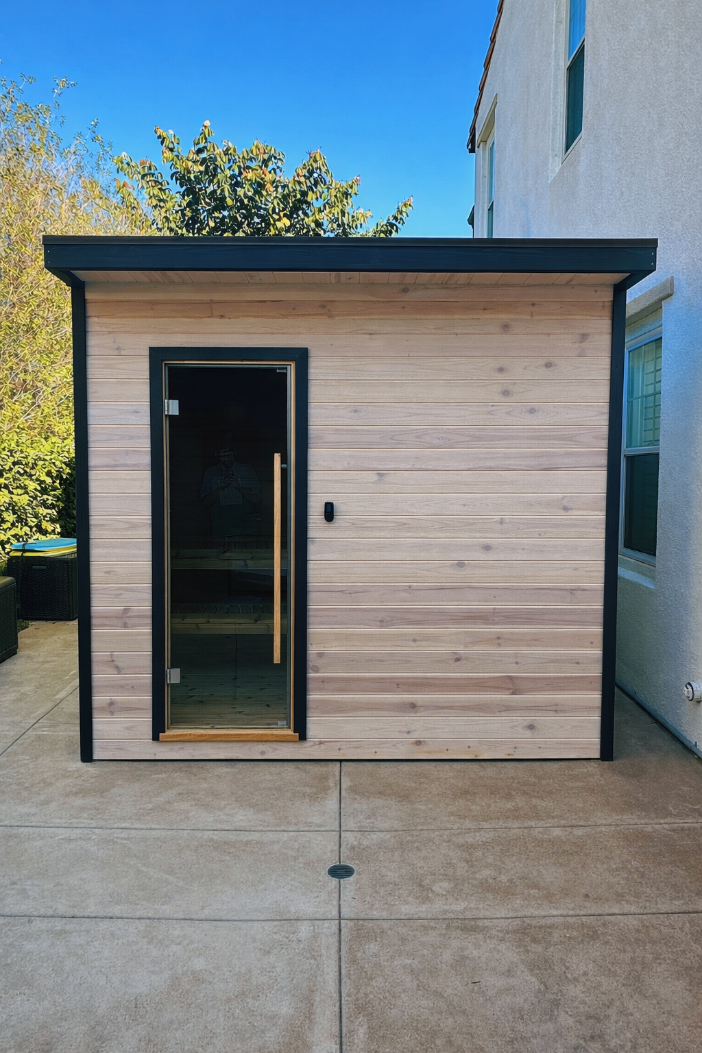 Small outdoor wooden sauna with glass door, black trim, and wooden handle, situated on a concrete patio next to a white building with window, with trees and blue sky in the background.