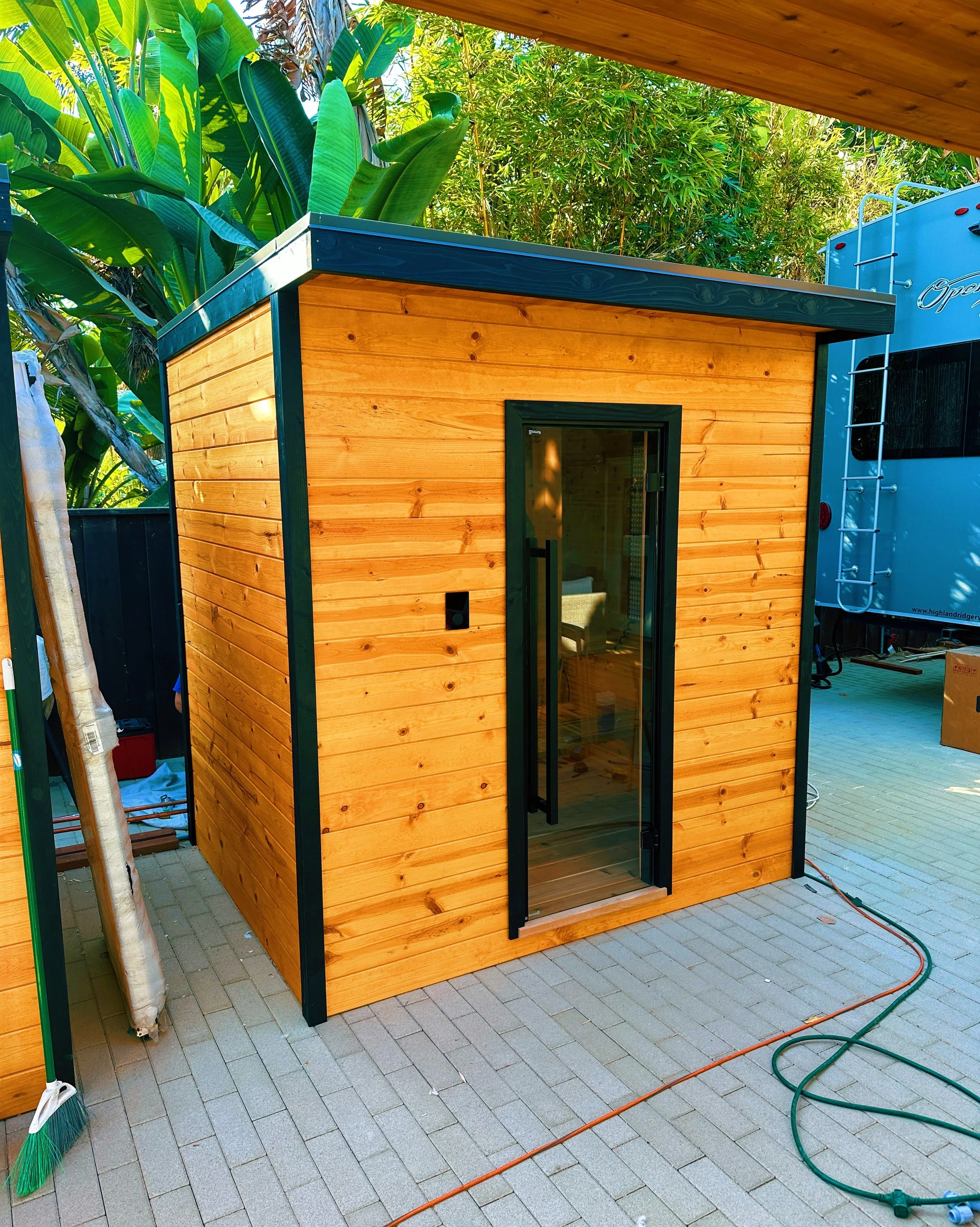 A small wooden shed with a black-framed glass door, sitting on a paved patio with green garden hose and broom nearby, surrounded by lush green tropical plants.