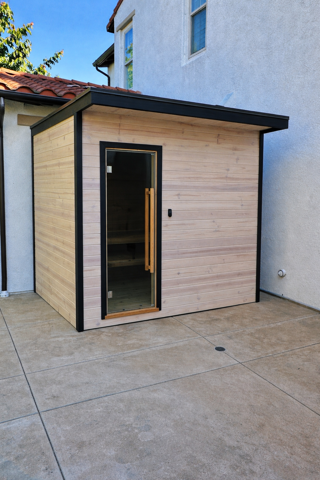 A small wooden sauna with a glass door and black trim, located on a concrete patio against the side of a house with light gray stucco walls.