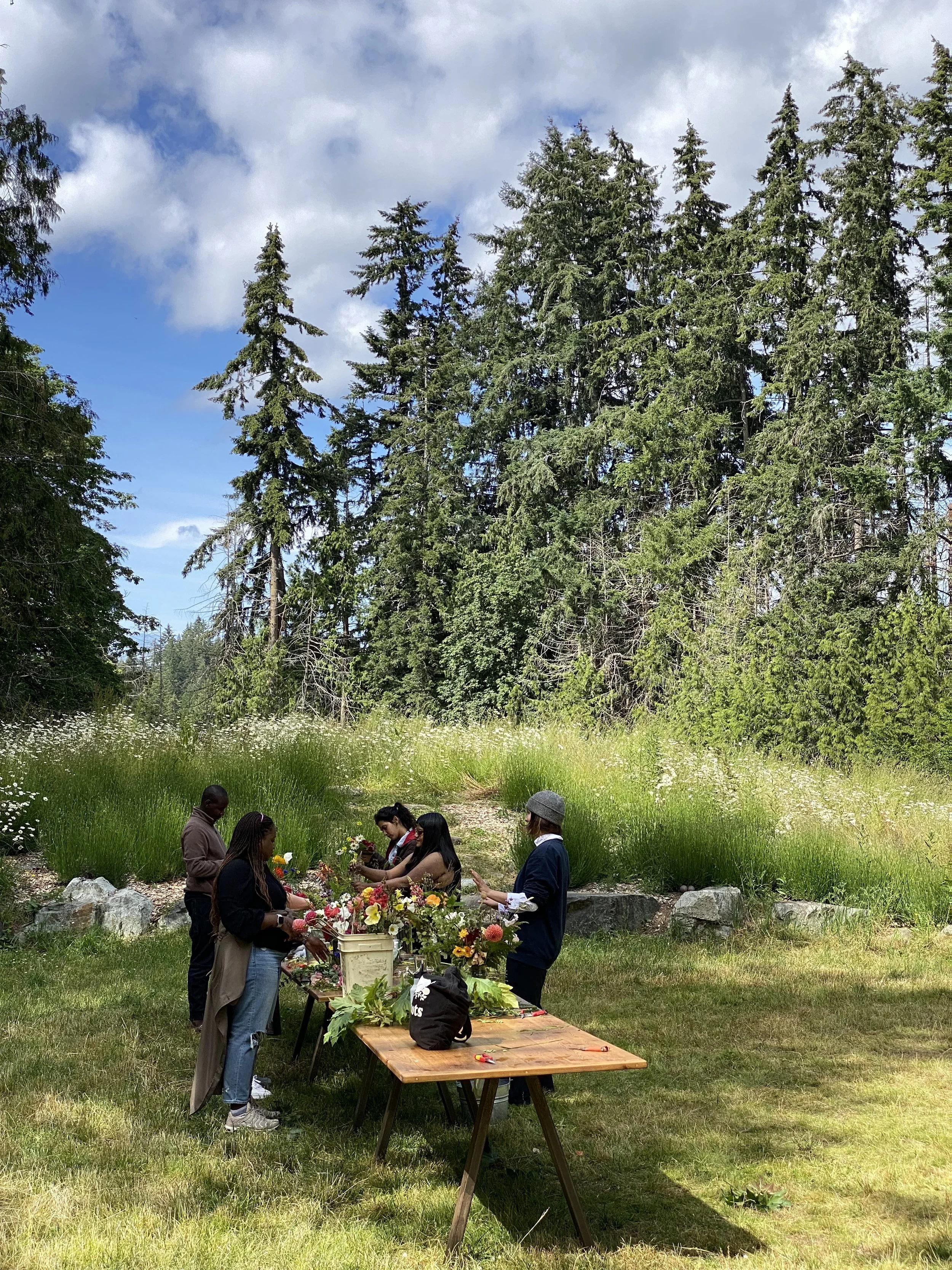 People gathered outdoors in a grassy area, arranging flowers on tables with tall trees and a partly cloudy sky in the background.