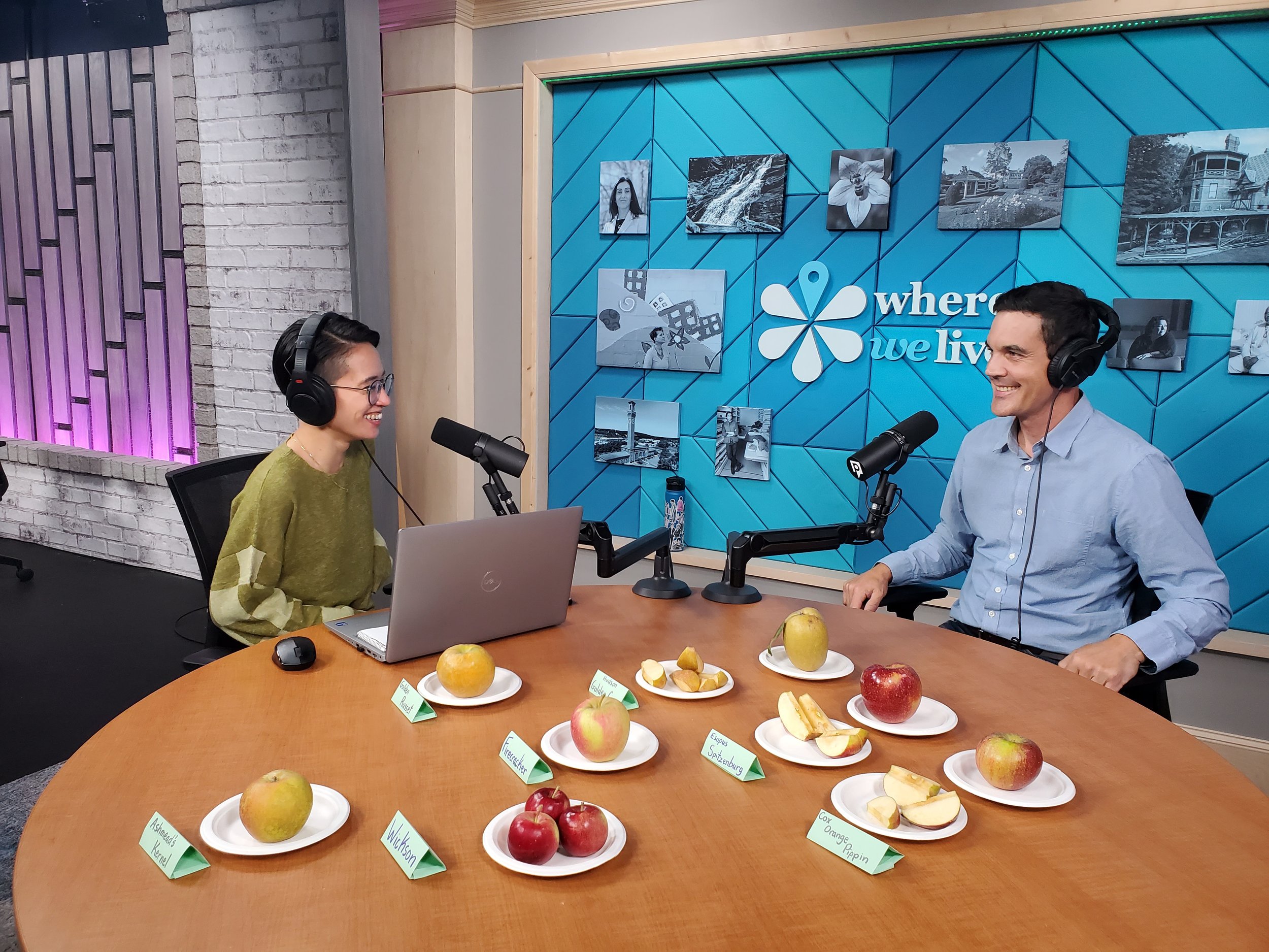 Two people, a woman and a man, speaking into microphones during a podcast or radio recording in a studio. They are smiling and wearing headphones. The studio has a blue wall with framed black and white photos and a sign that reads 'where we live.' A table in front of them displays various apples and fruit samples, each labeled with small tags.