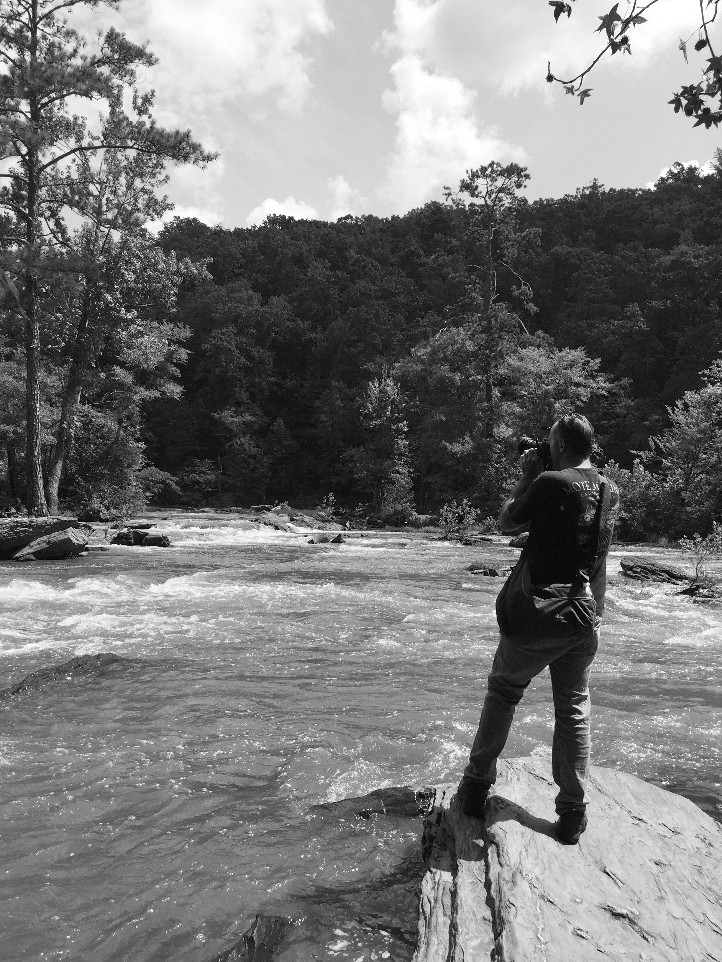 Charles Titterington standing on a rock, photographing a rushing river surrounded by forest.