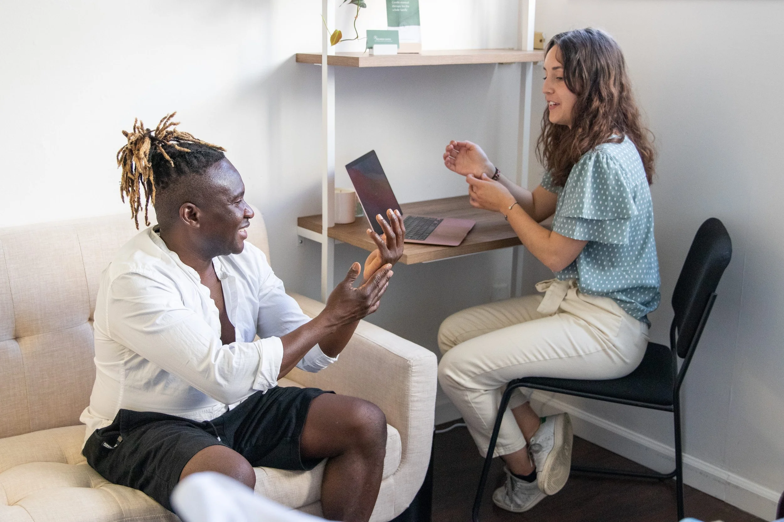 A woman with dark skin, light brown hair, wearing a blue polka dot shirt and beige pants, sitting on a chair with folded arms, smiling and talking to a man with dark skin, blonde dreadlocks, wearing a white shirt and black shorts, sitting on a beige sofa, with a laptop on the wooden shelf in the background.
